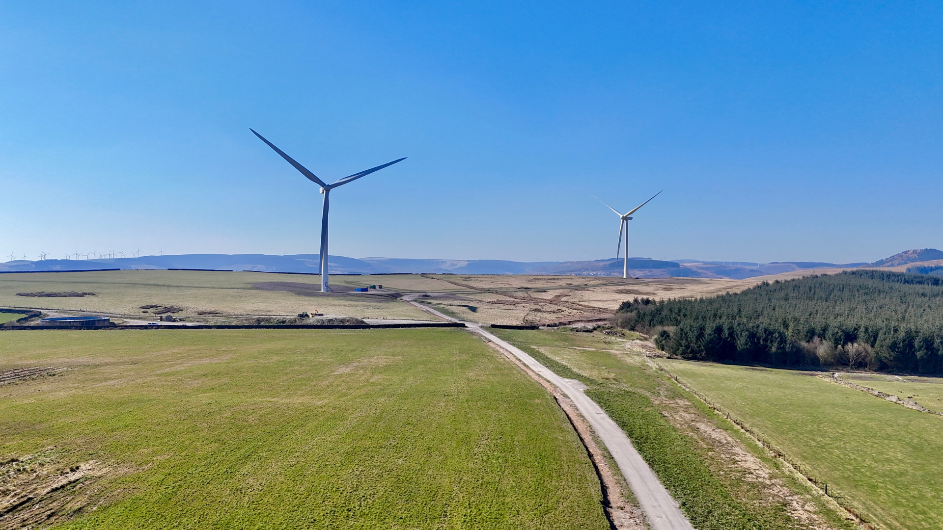A landscape with rolling green fields, wind turbines, and a clear blue sky.