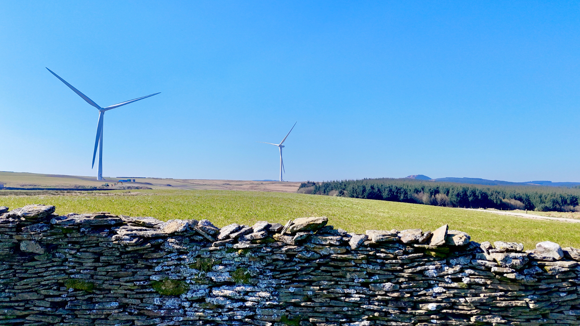 Wind turbines in a rural landscape with a stone wall in the foreground, green fields, and a clear blue sky.
