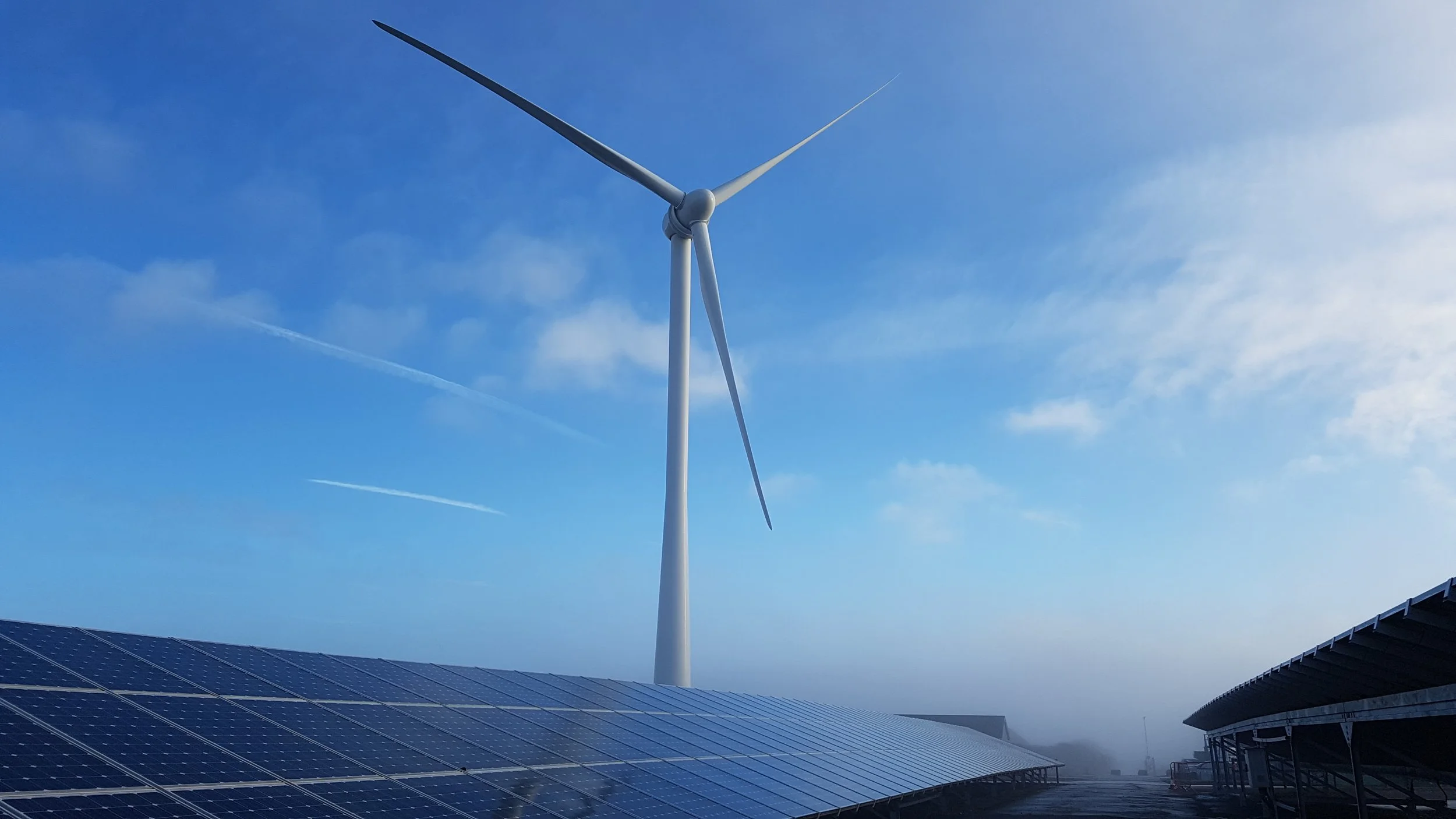 Solar panels on a rooftop with a wind turbine in the background under a partly cloudy sky.