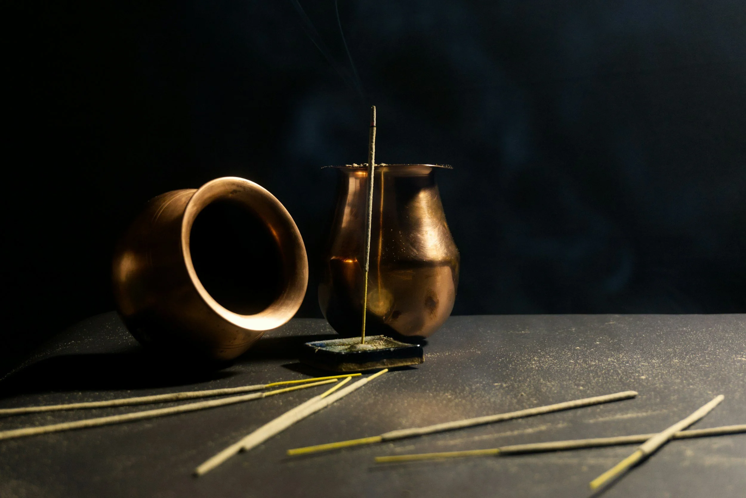 A still life photograph featuring two metallic copper incense holders, one on its side and one standing upright, with incense sticks and ashes on a dark surface.