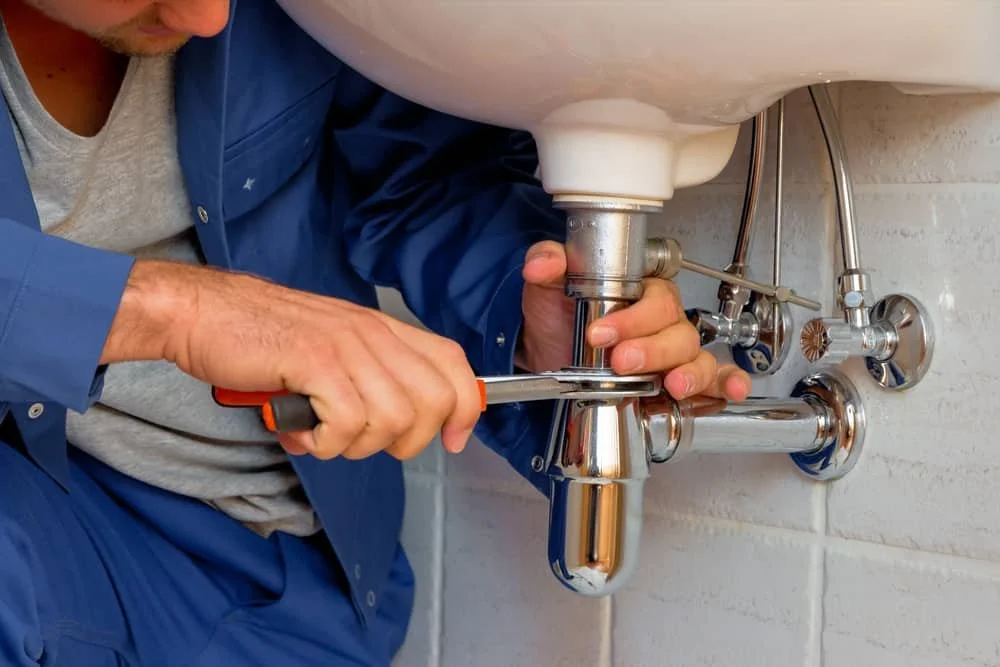 A person tightening a pipe fitting underneath a bathroom sink with a screwdriver.