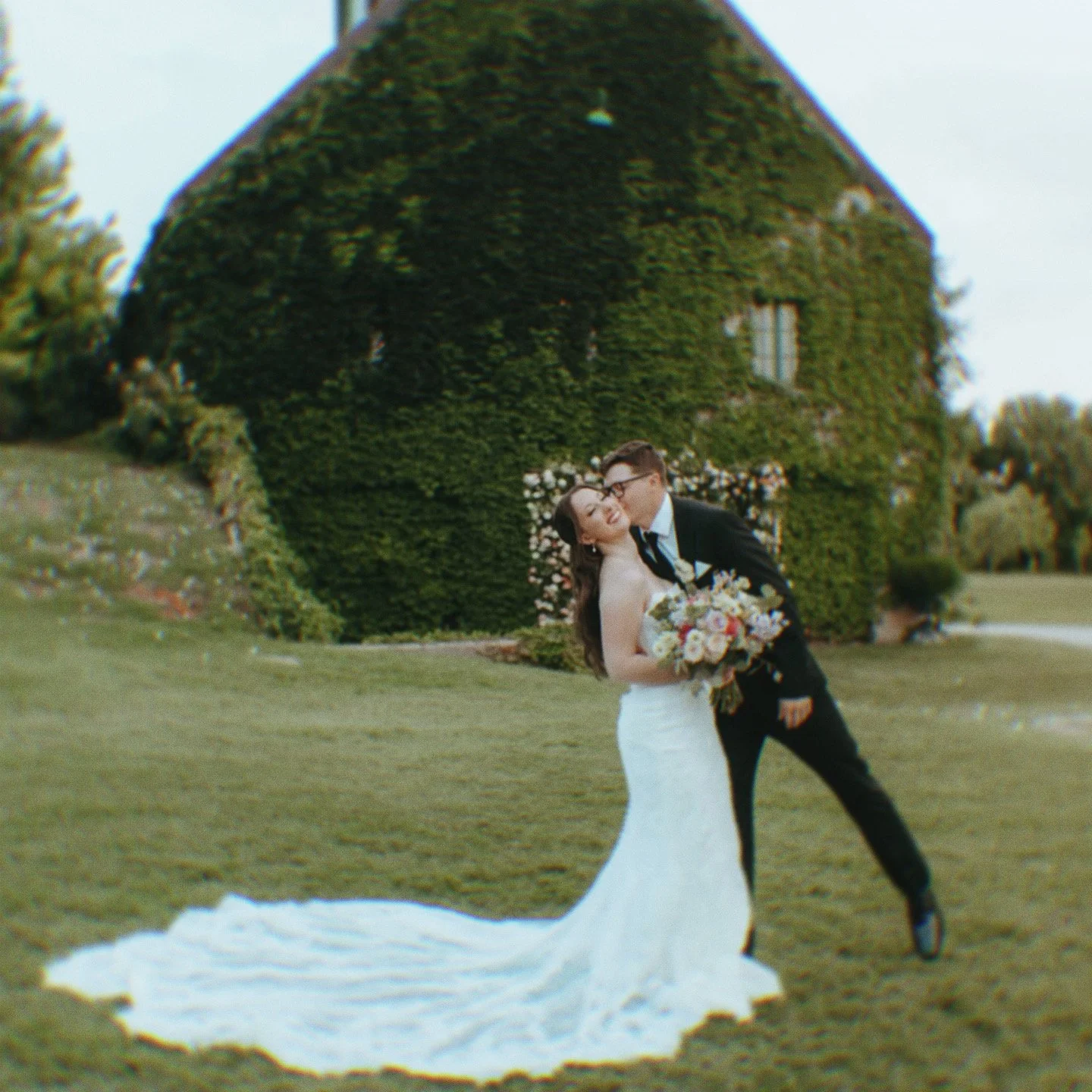 A newlywed couple sharing a kiss outdoors with a green ivy-covered building in the background, the bride holding a bouquet of flowers, and the groom leaning in to kiss her.