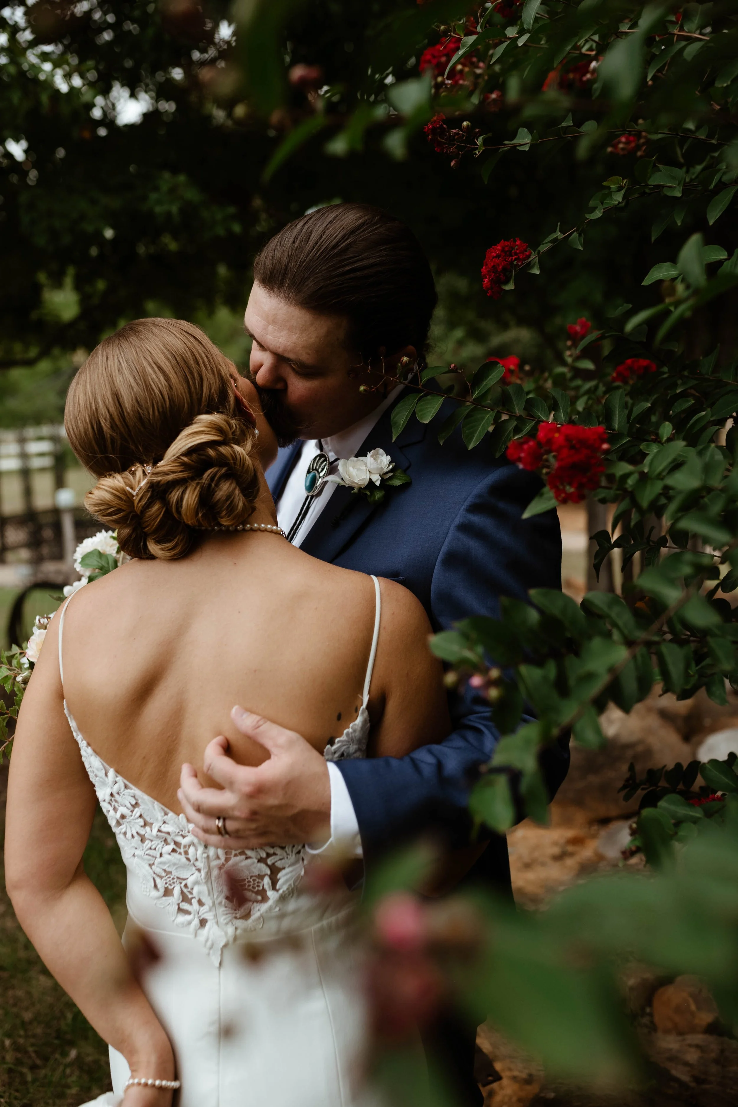 A bride and groom sharing a kiss outdoors surrounded by green foliage and red flowers.