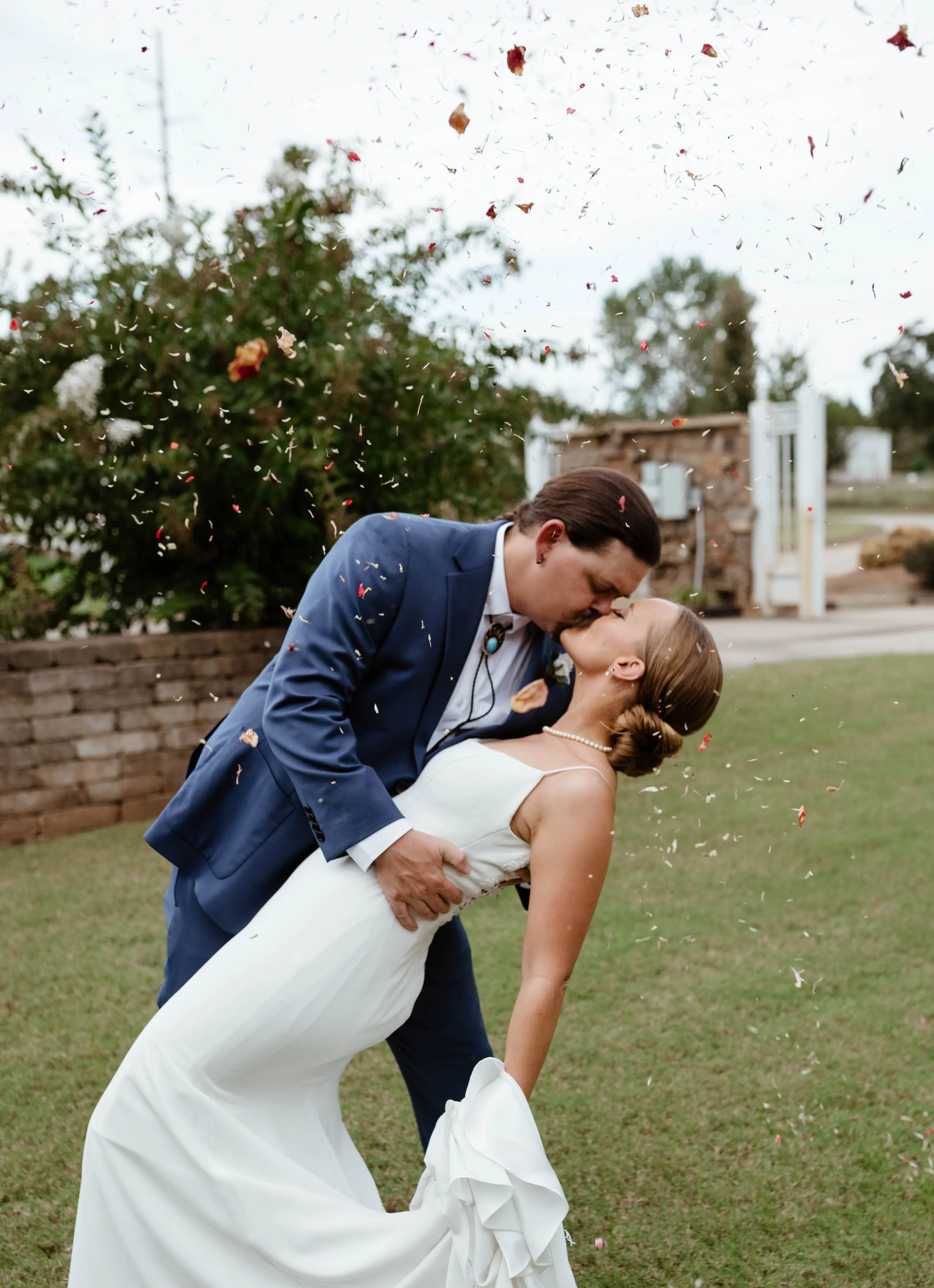 A couple in wedding attire sharing a kiss outdoors, with confetti falling around them.