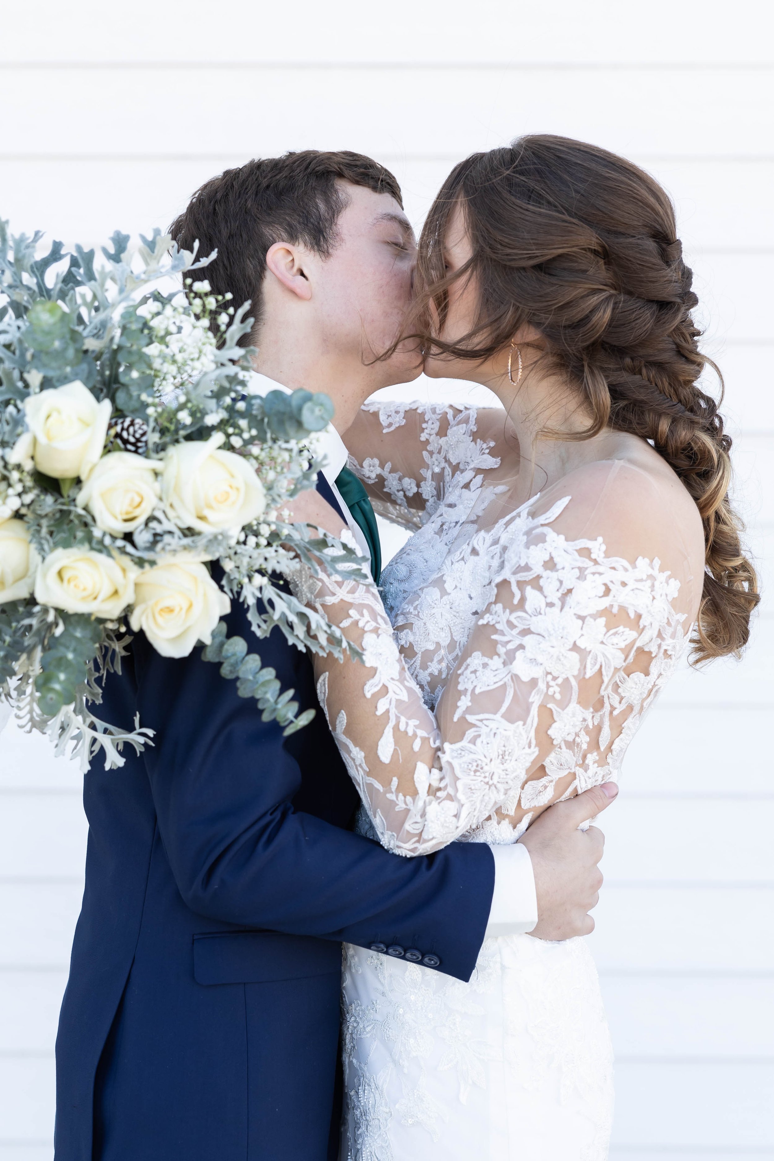 A bride and groom kissing, with the bride holding a bouquet of white roses and greenery.