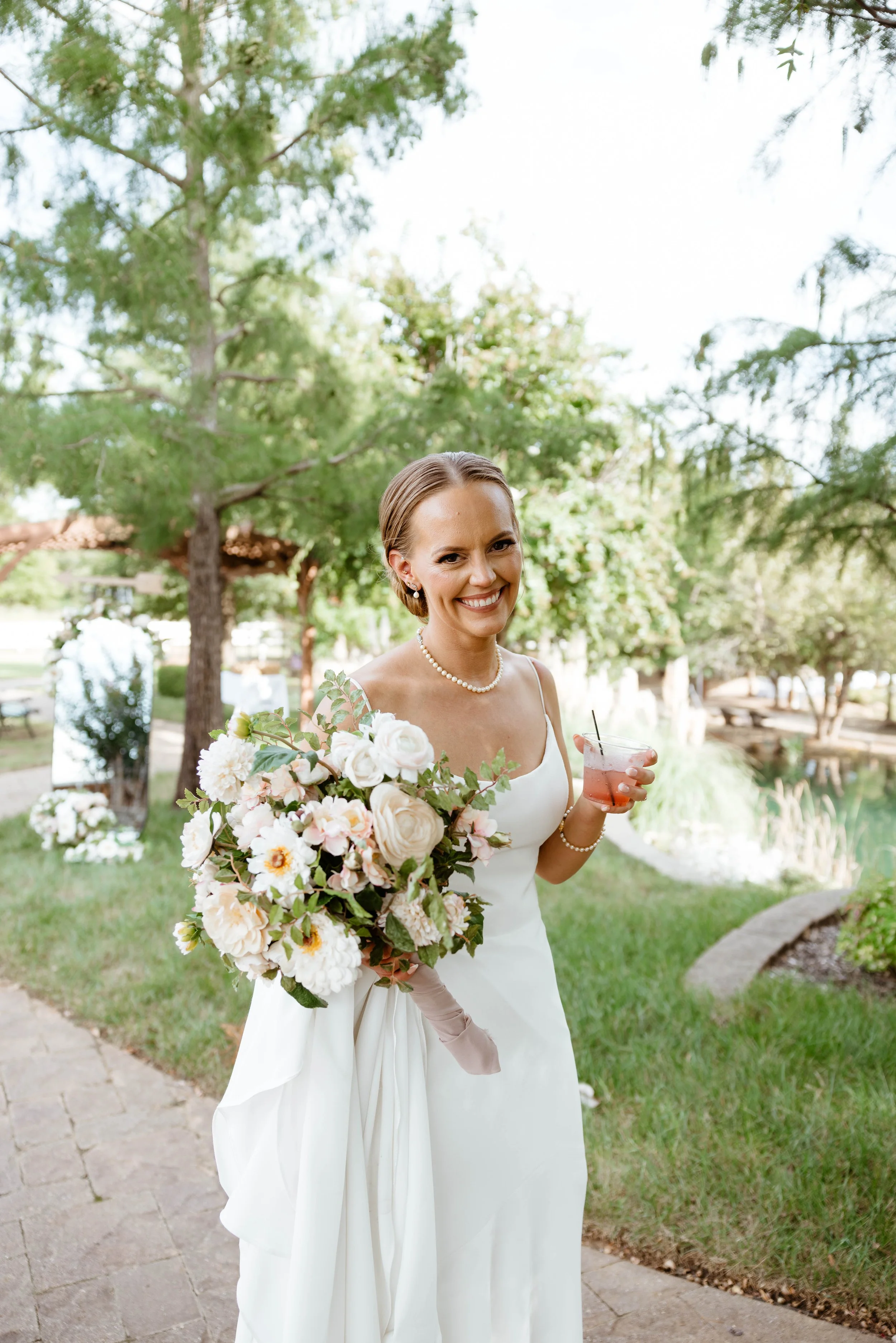 A smiling woman in a white dress holding a bouquet of white and blush flowers and a pink cocktail, standing outdoors near a pond with trees and grass.