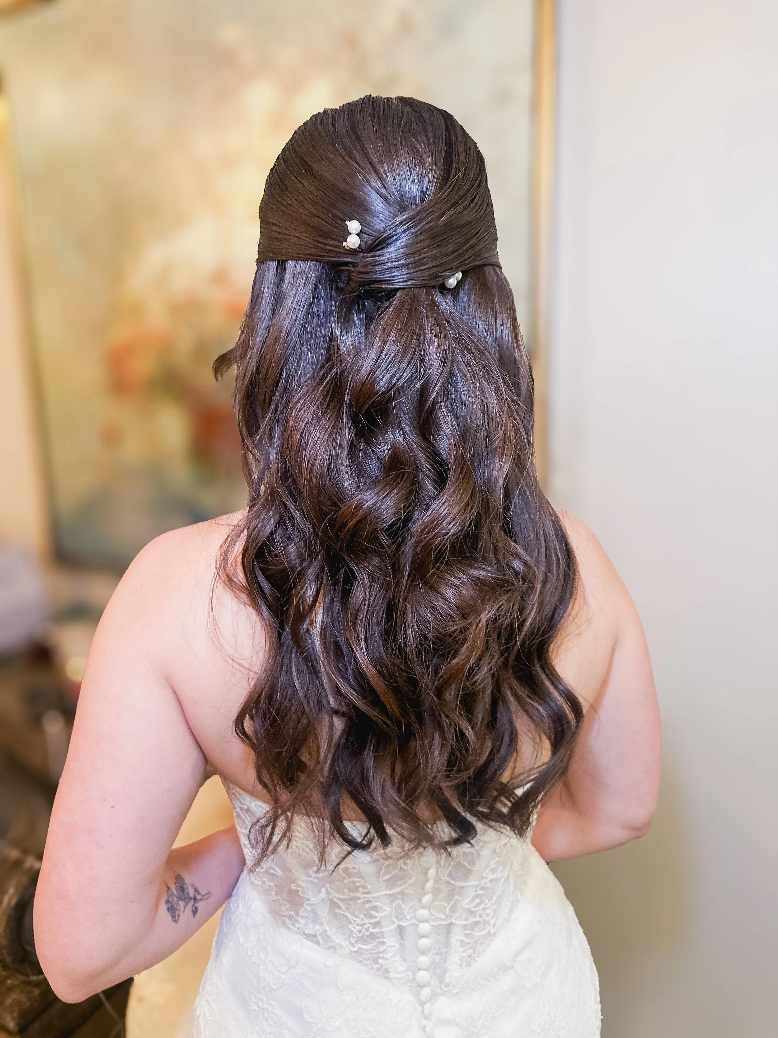 Back view of a woman with long, wavy brown hair styled with a half-updo decorated with pearl pins, wearing a white lace dress.