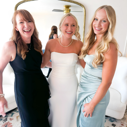 Three women dressed in formal attire standing together indoors, smiling, with a mirror and ceiling fan in the background.