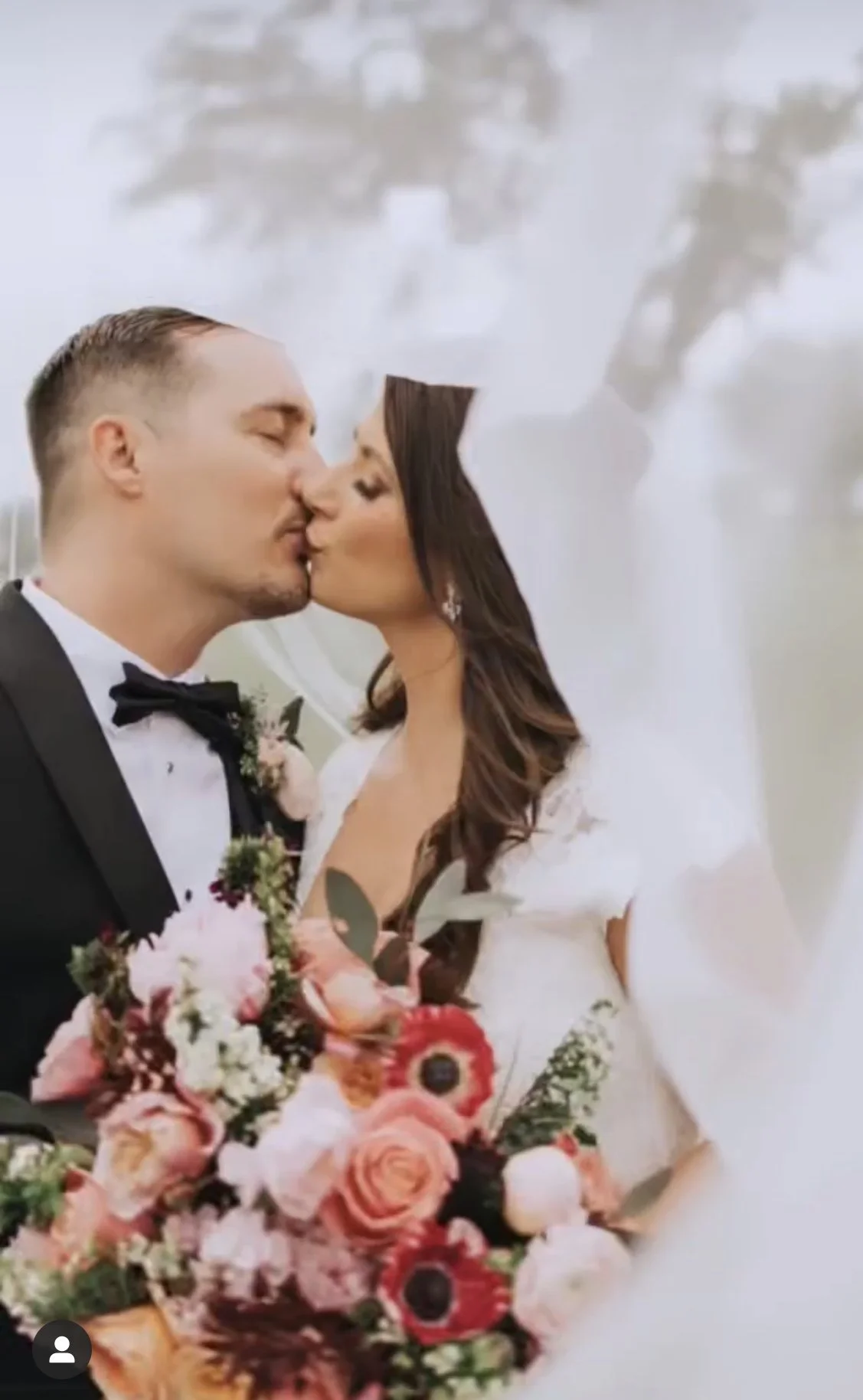 A bride and groom share a kiss on their wedding day, with the bride holding a large bouquet of flowers.
