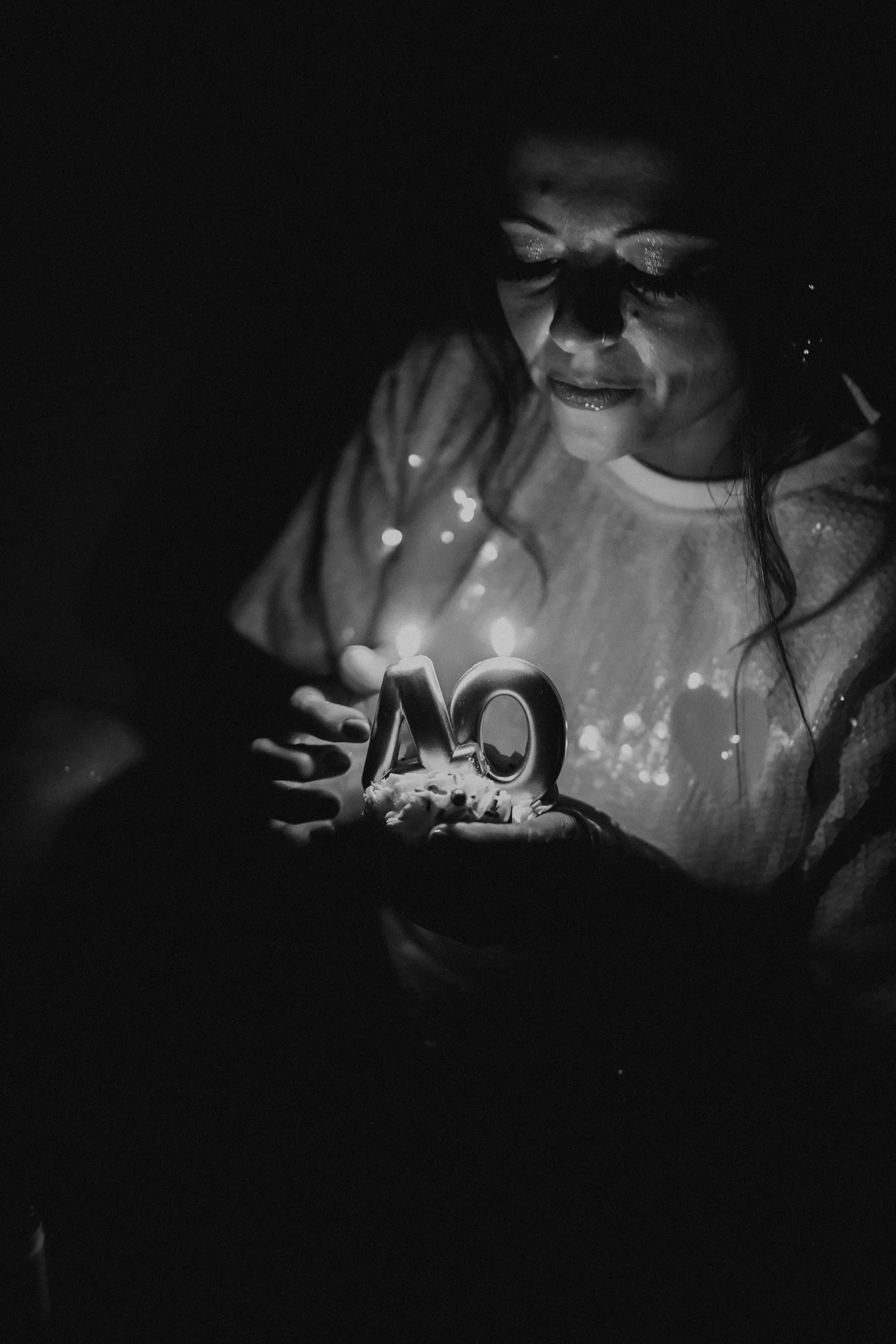 A woman in a glittery sweater holding a cupcake with lit candles and the number 40, celebrating her 40th birthday in a dark environment.