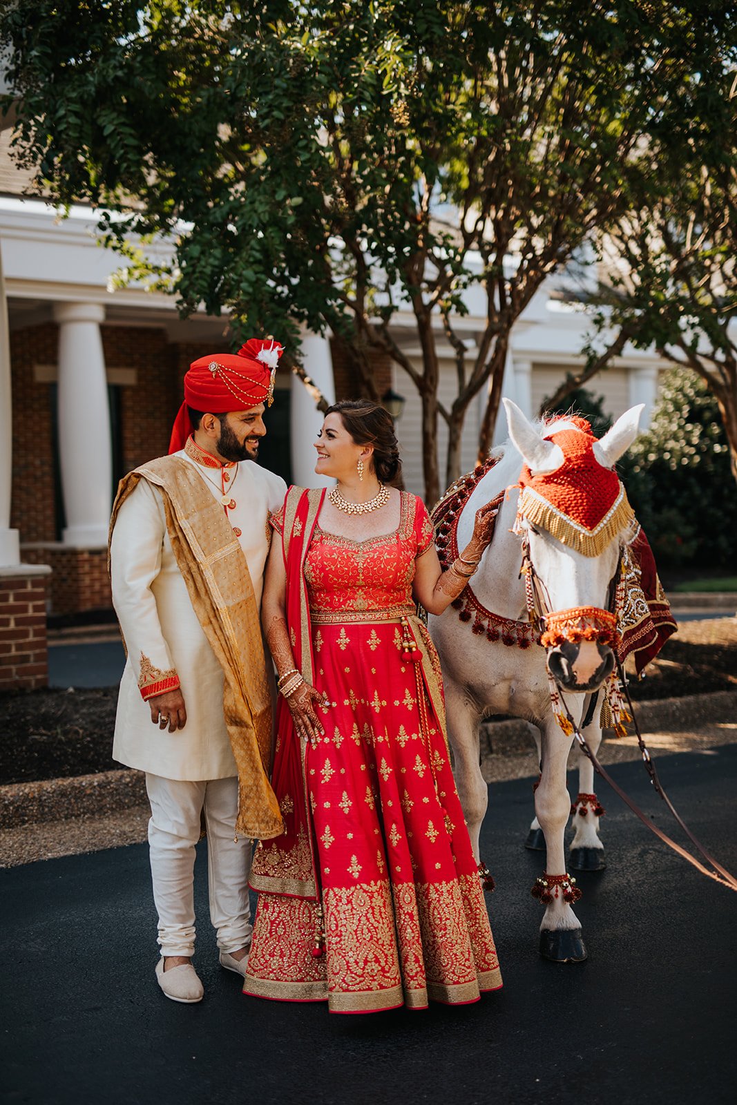 A bride and groom in traditional Indian wedding attire standing with a decorated white horse, outdoors with trees and a building in the background.