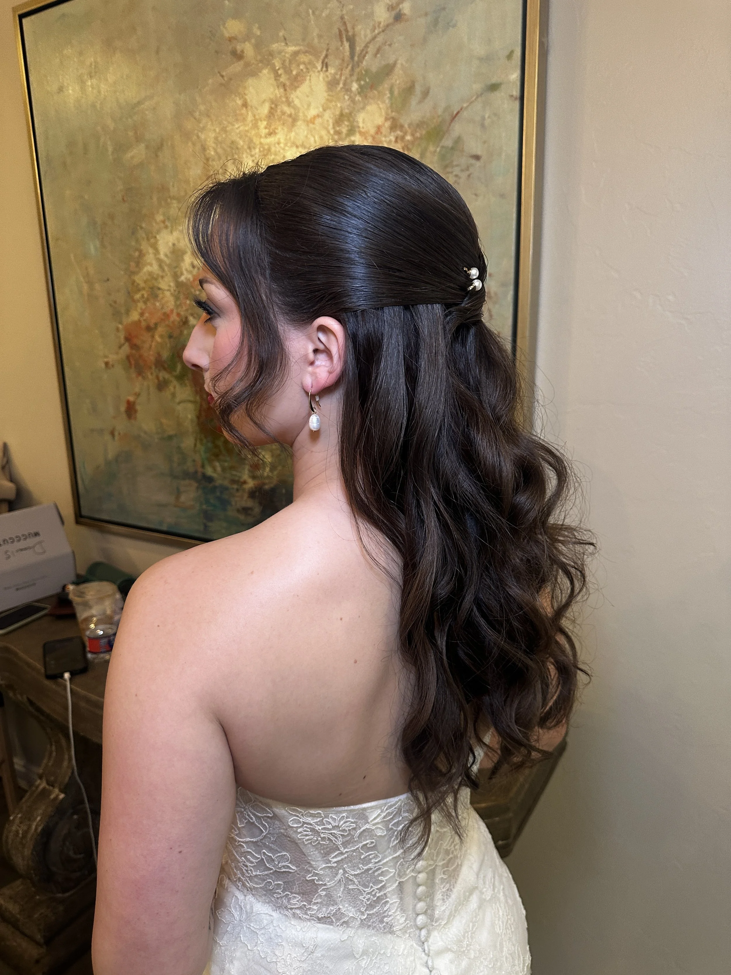 A woman with dark, wavy hair styled in loose curls, adorned with pearl hairpins, wearing pearl drop earrings and a strapless white lace dress, standing indoors against a textured wall and abstract painting.