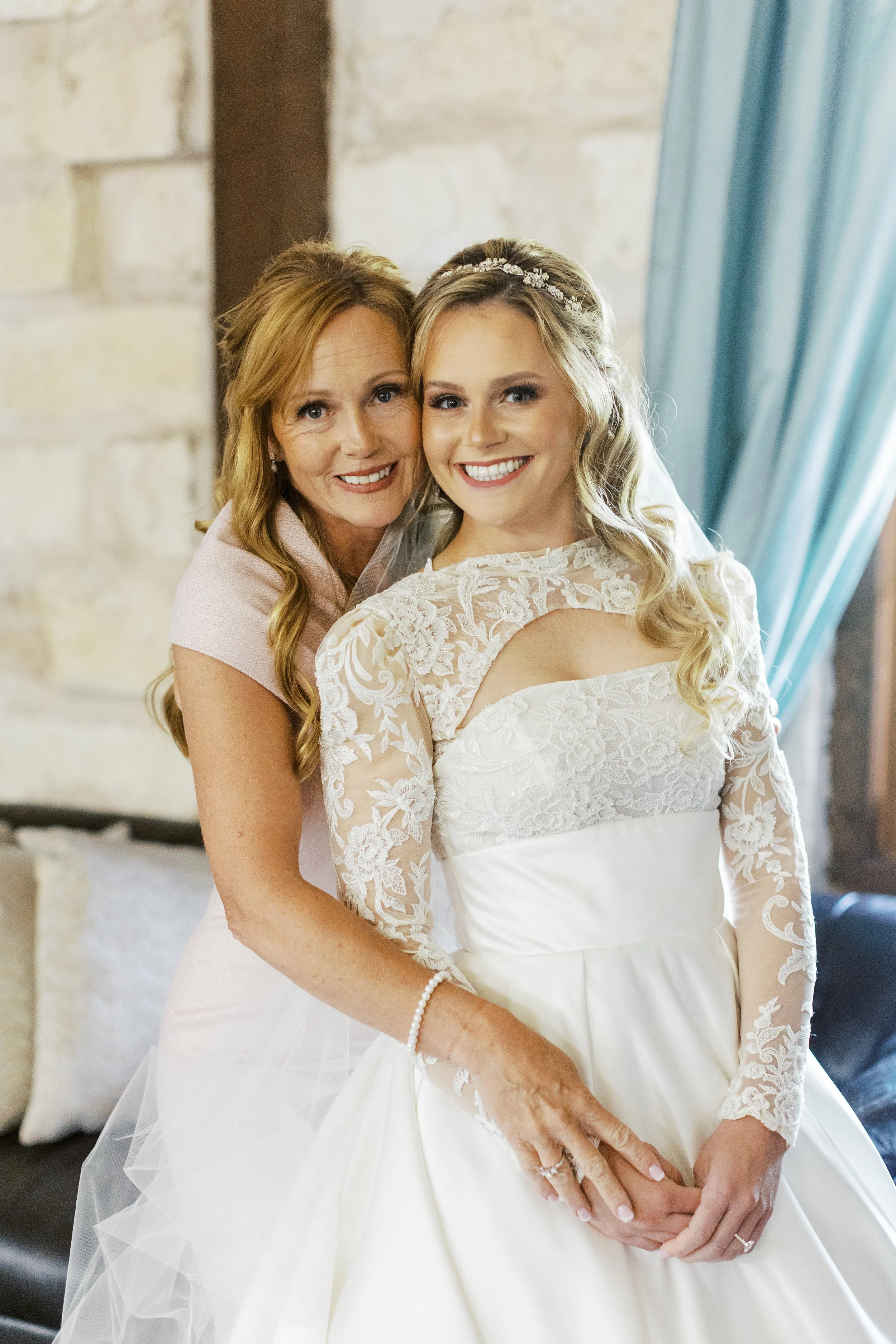 A bride in a white lace wedding gown with a sweetheart neckline and long lace sleeves, smiling, with an older woman, possibly her mother, hugging her from behind, both smiling, in a room with stone walls and blue curtains.