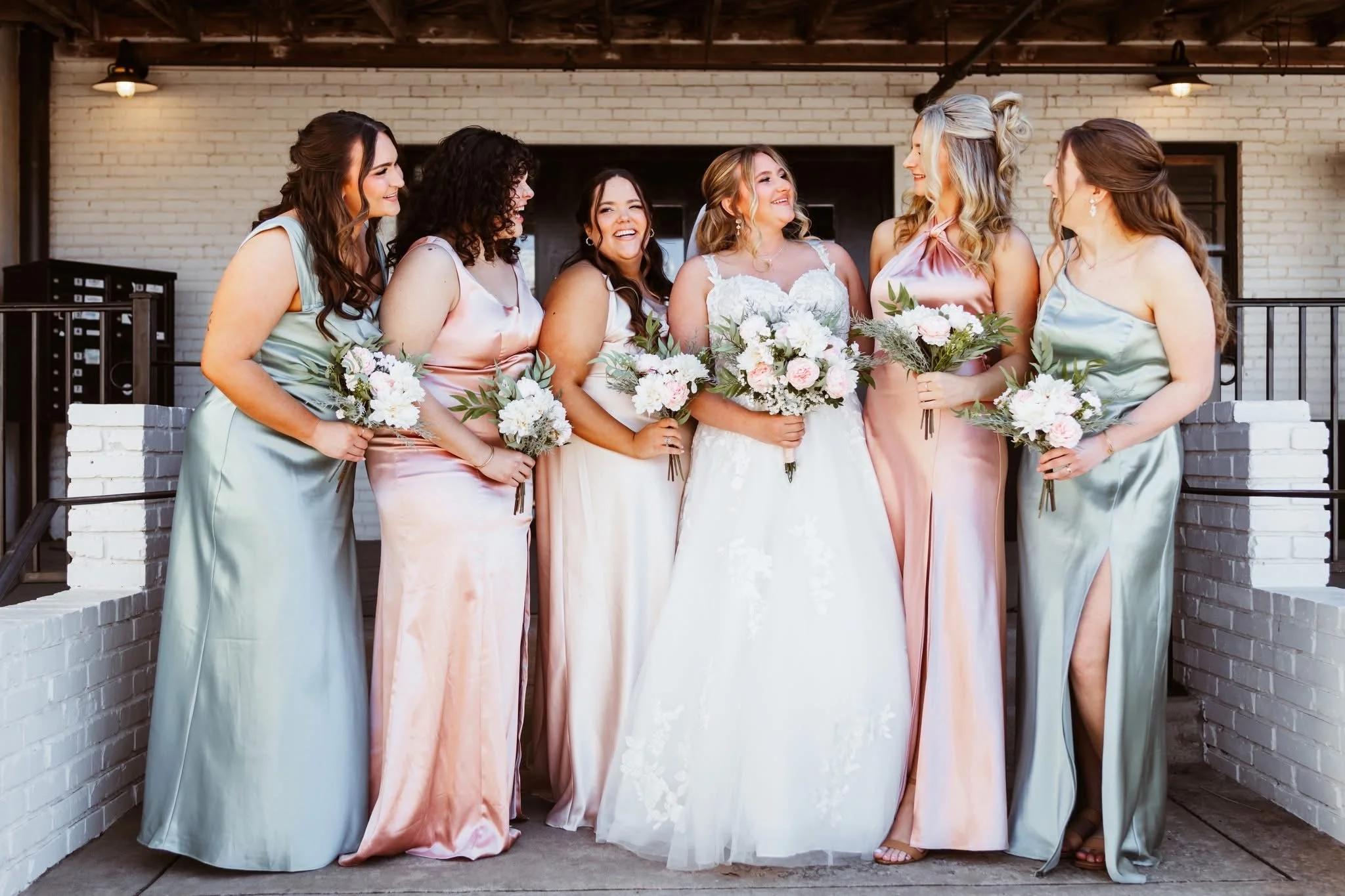 Group of six women dressed in pastel wedding attire, holding bouquets, standing on a patio in front of a white brick wall.