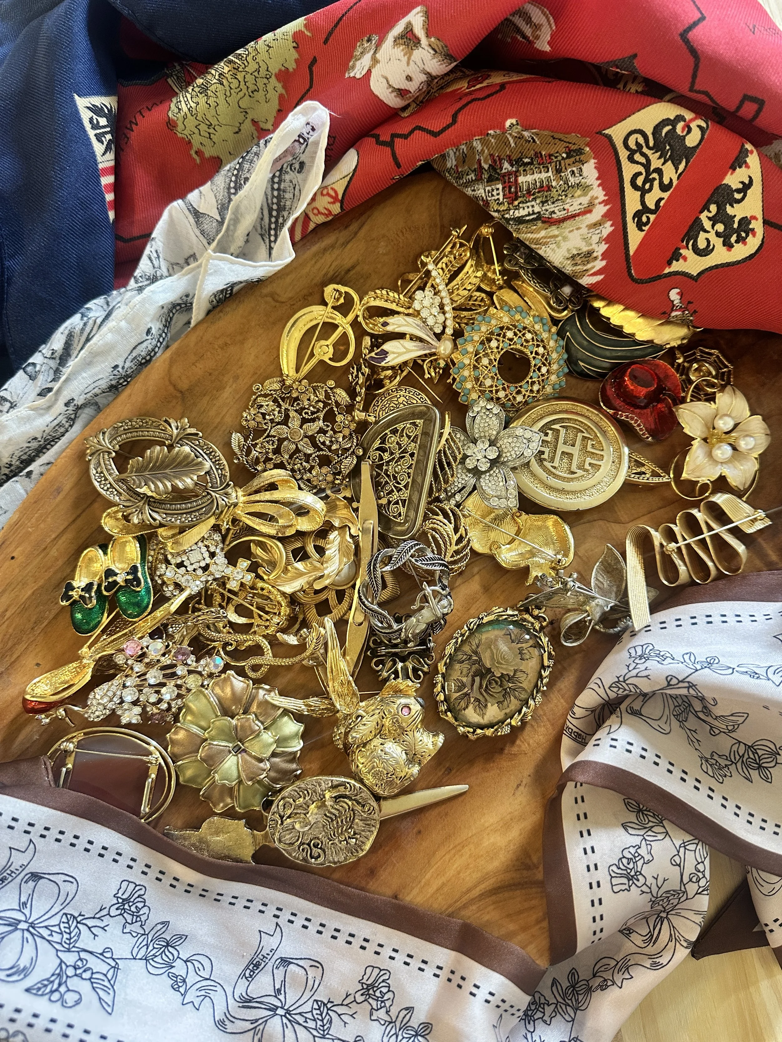 A variety of gold and silver jewelry including necklaces, bracelets, earrings, and brooches, scattered on a wooden surface surrounded by colorful cloths.