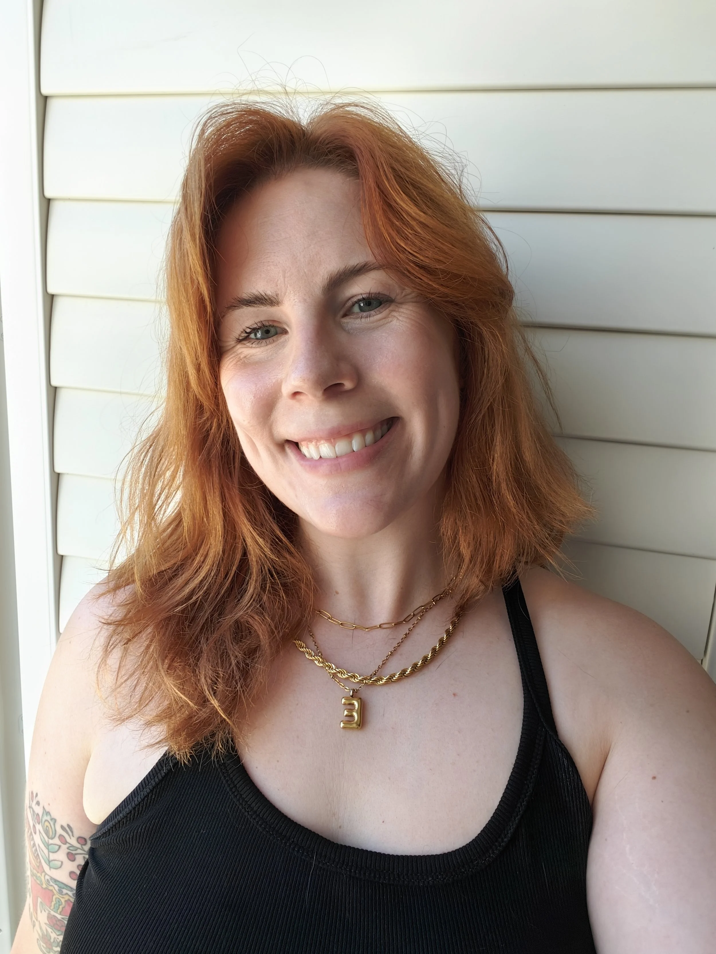 Close-up of a smiling woman with red hair, tattoos on her arm, wearing a black top and gold jewelry, in front of white siding.