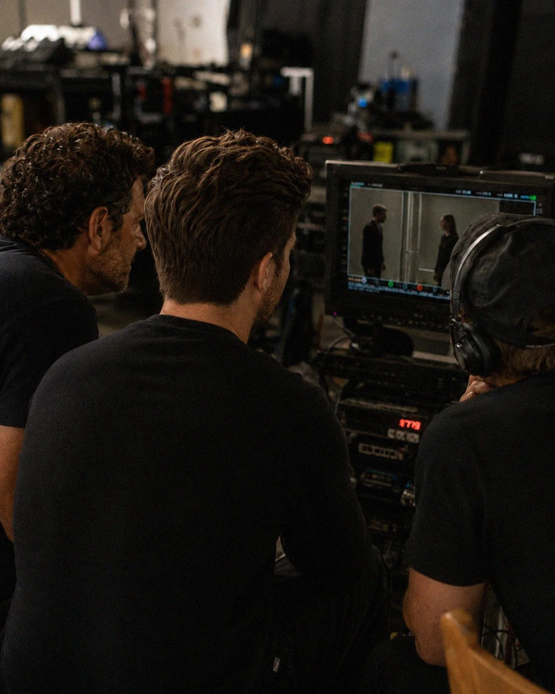 Three men on a film set watch a scene on a monitor, with one man wearing headphones.