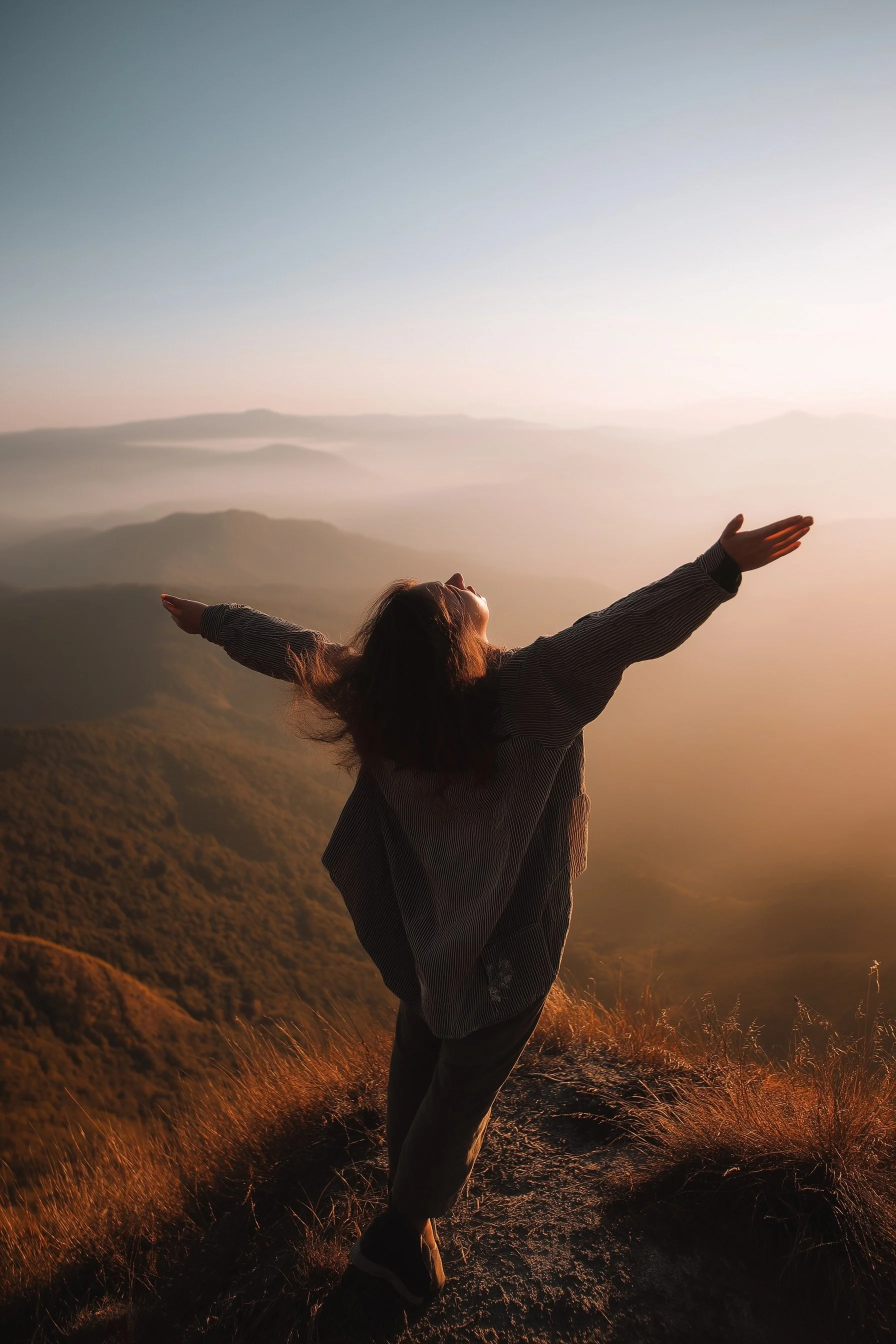Person standing with arms outstretched on mountain top during sunrise, overlooking hills and valleys.