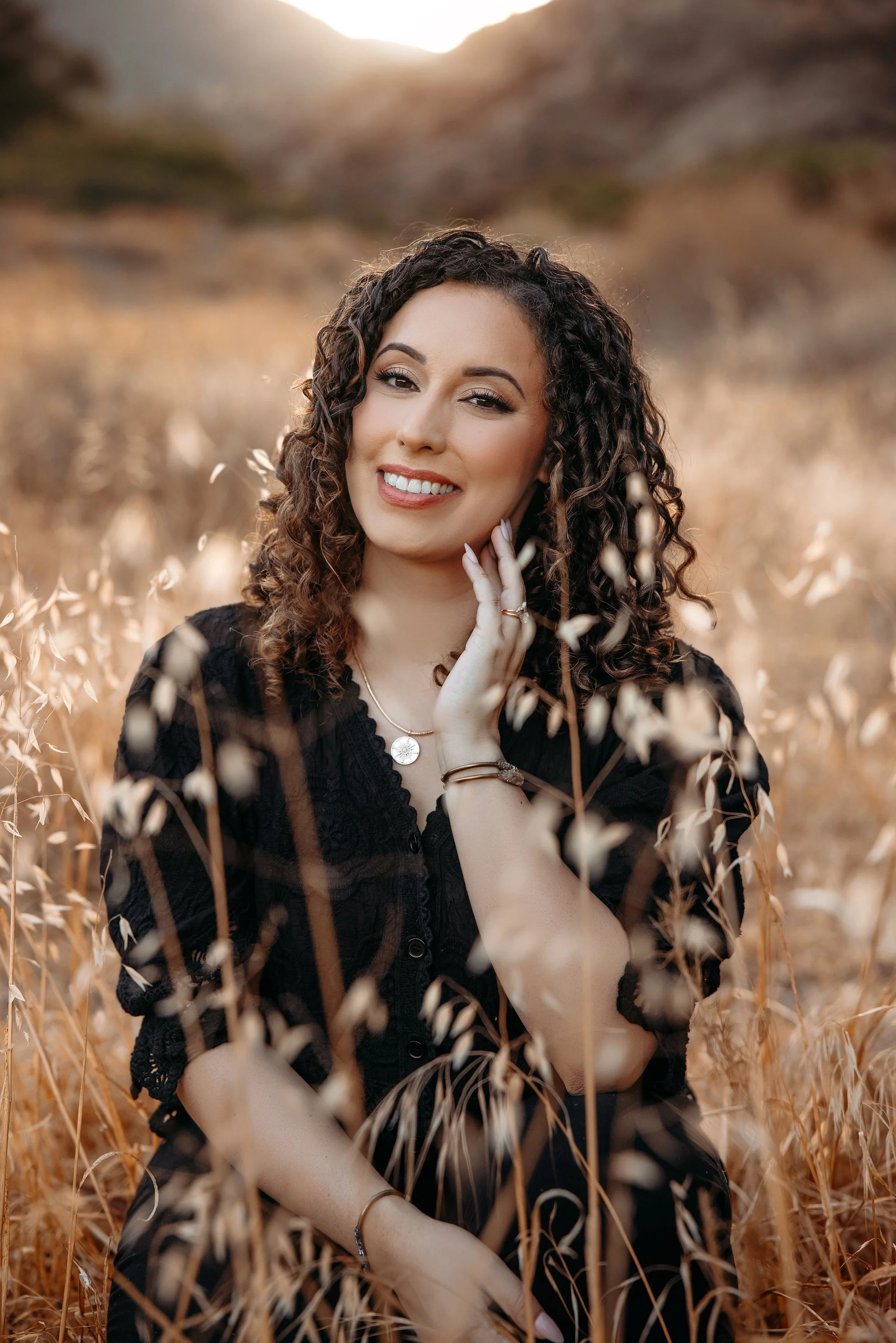 A woman with curly hair smiling, sitting in a field of tall, golden grass with mountains in the background, during sunset.