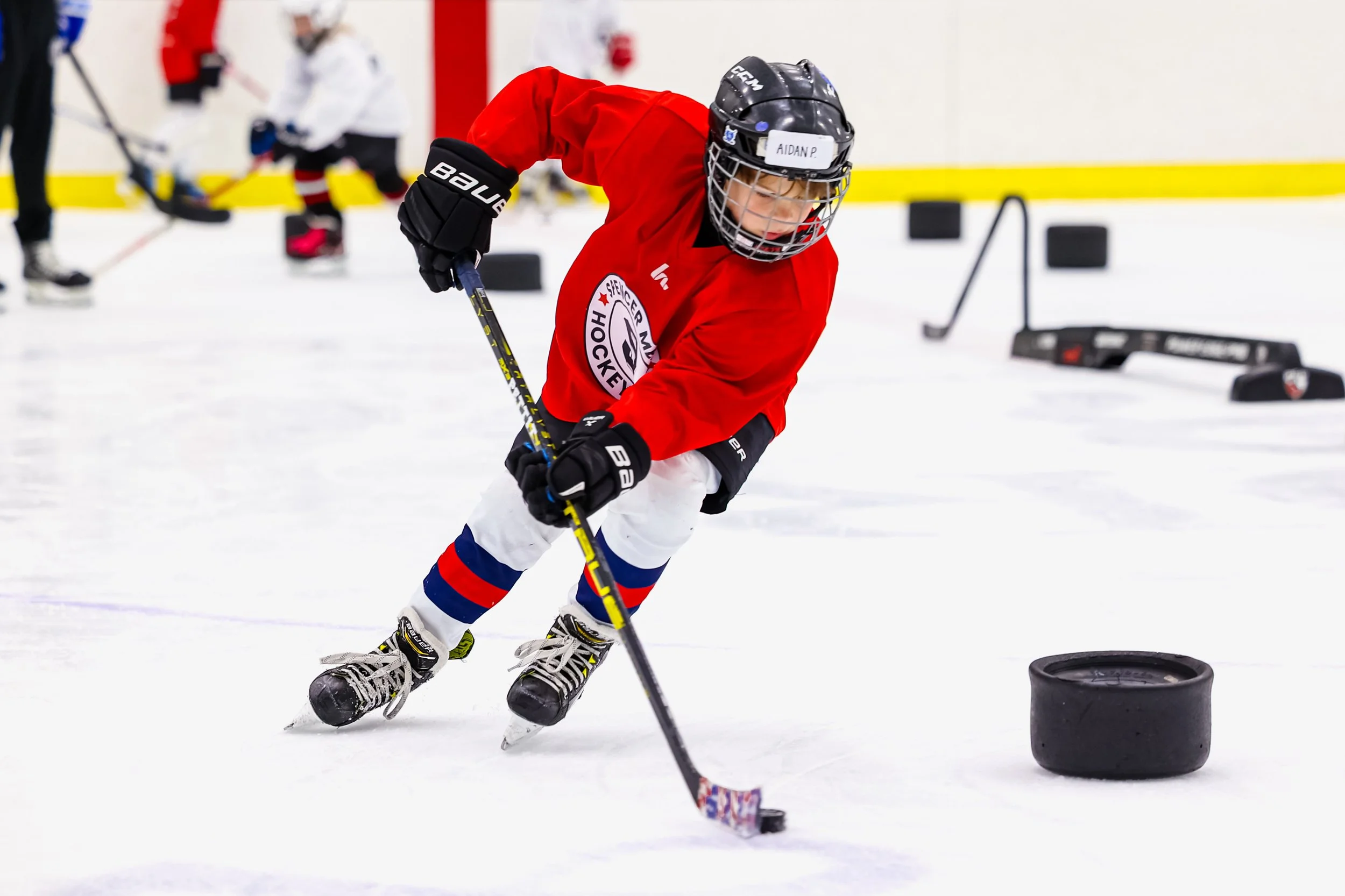 Young hockey player in red jersey practicing on ice rink with a hockey stick near a hockey puck, wearing helmet and gloves, with other players and equipment in the background.