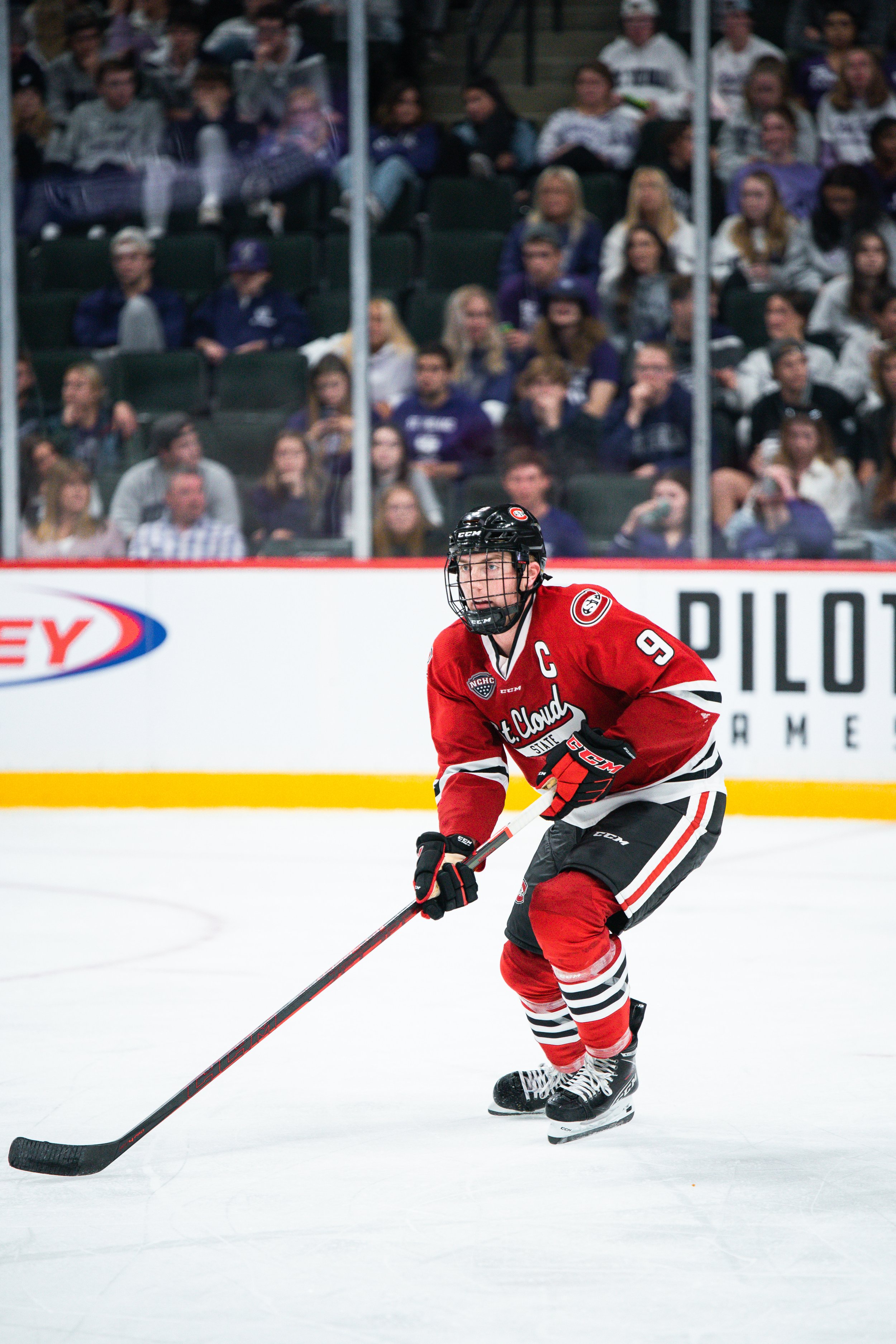 Hockey player in red jersey with 'C' on their chest, wearing helmet, gloves, and skates, skating on ice rink during a game, with spectators watching in the background.