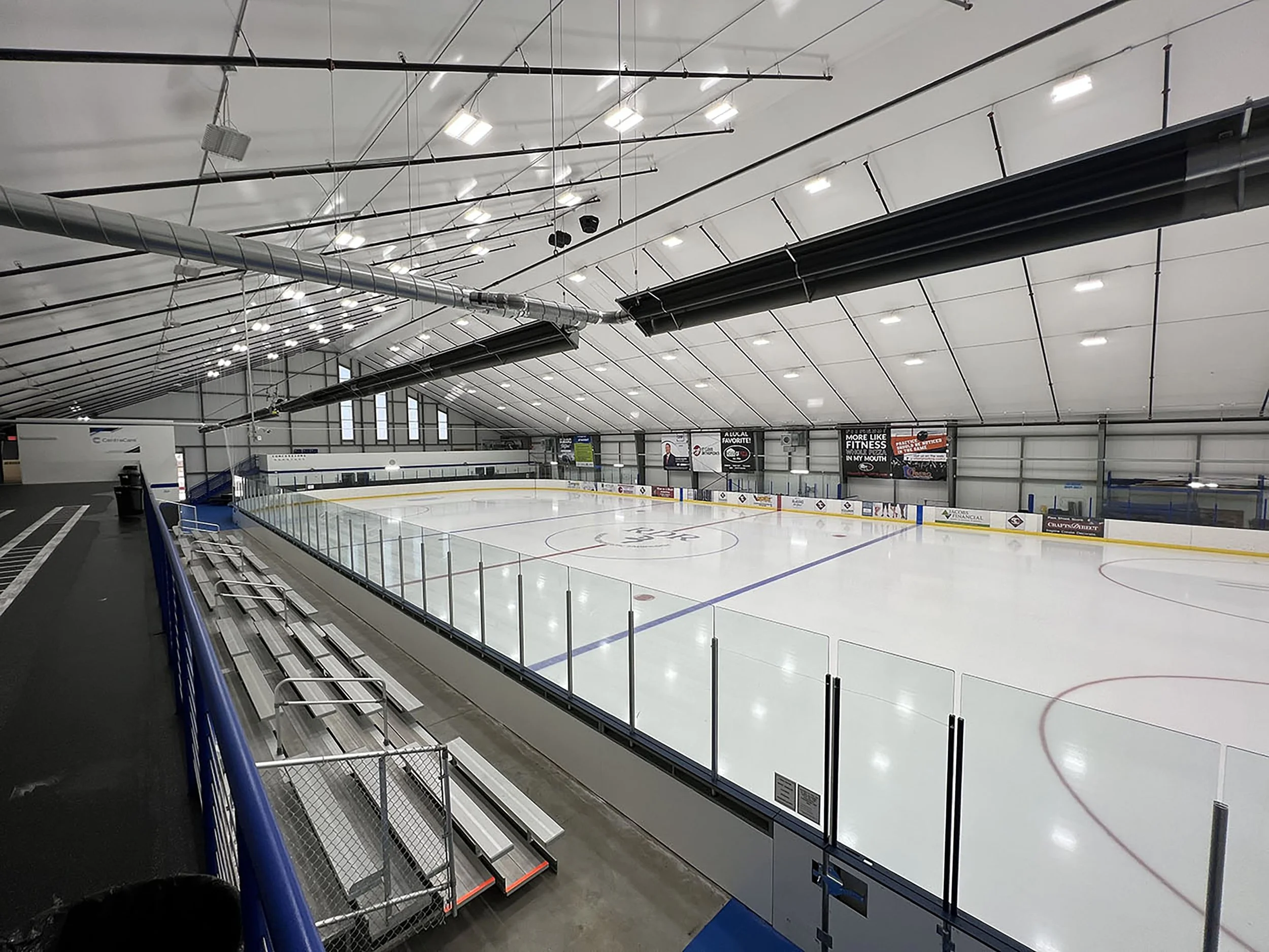An empty indoor ice hockey rink with white ice, marked with red and blue lines, surrounded by clear protective glass and padded boards. Bleacher seats are along one side, and signs and digital screens are on the far wall under a high curved ceiling.