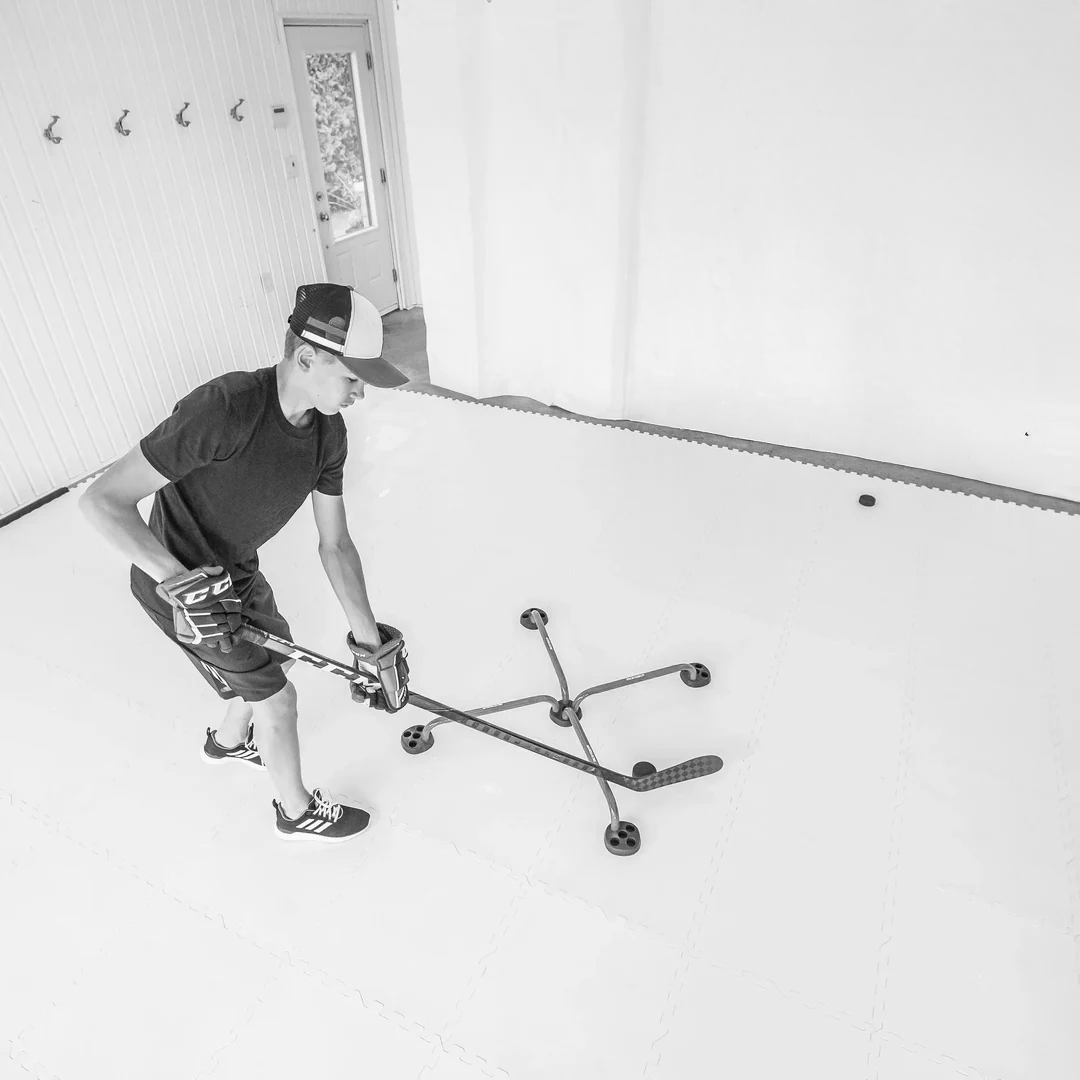 A young man in casual clothing and a cap is playing hockey indoors on a white floor, using a hockey stick and a puck. The room has a white wall, hooks, and a door with a window.