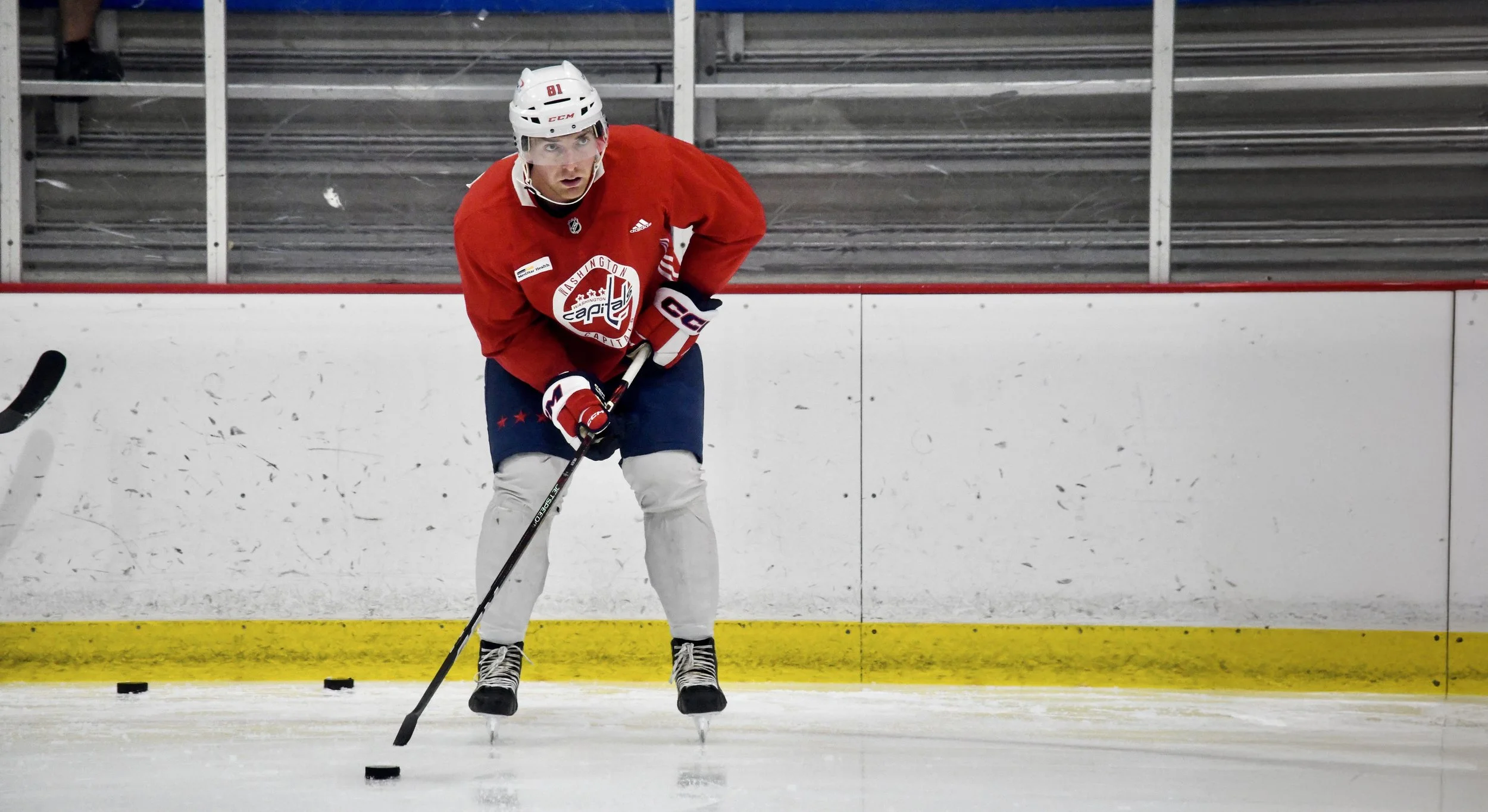 An ice hockey player in a red jersey, white helmet, and white pants, practicing on an indoor rink.