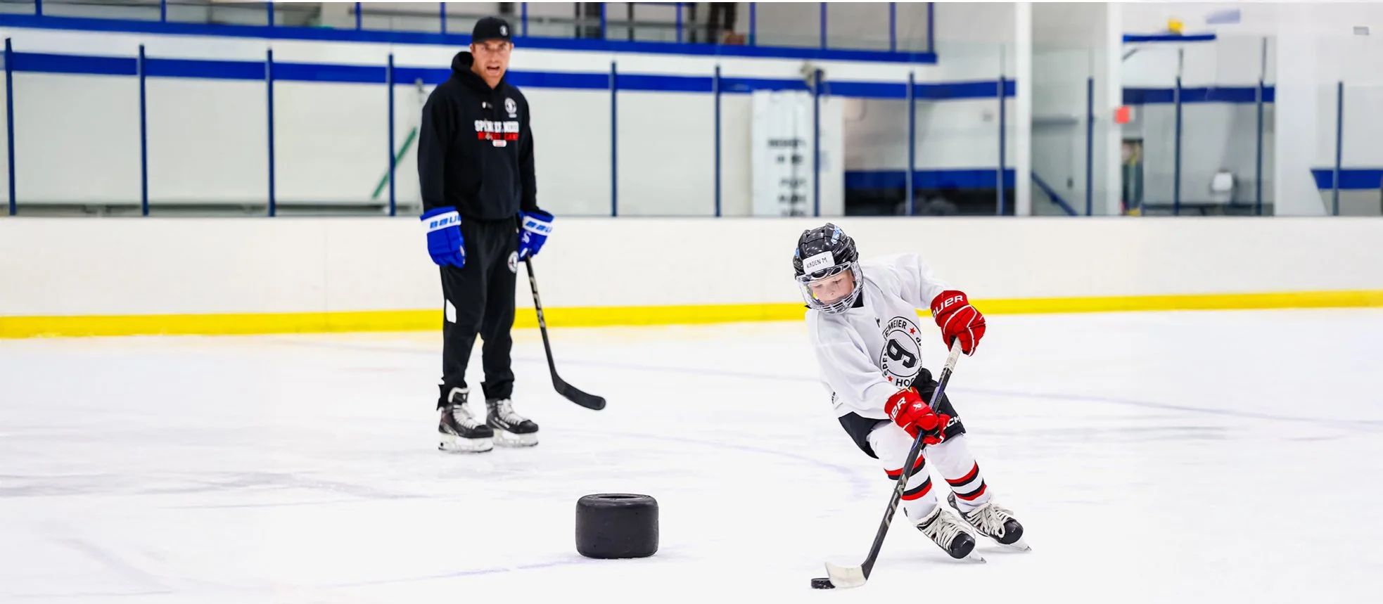 A young ice hockey player in a white jersey and red gloves practices puck handling with a coach observing on an indoor ice rink.