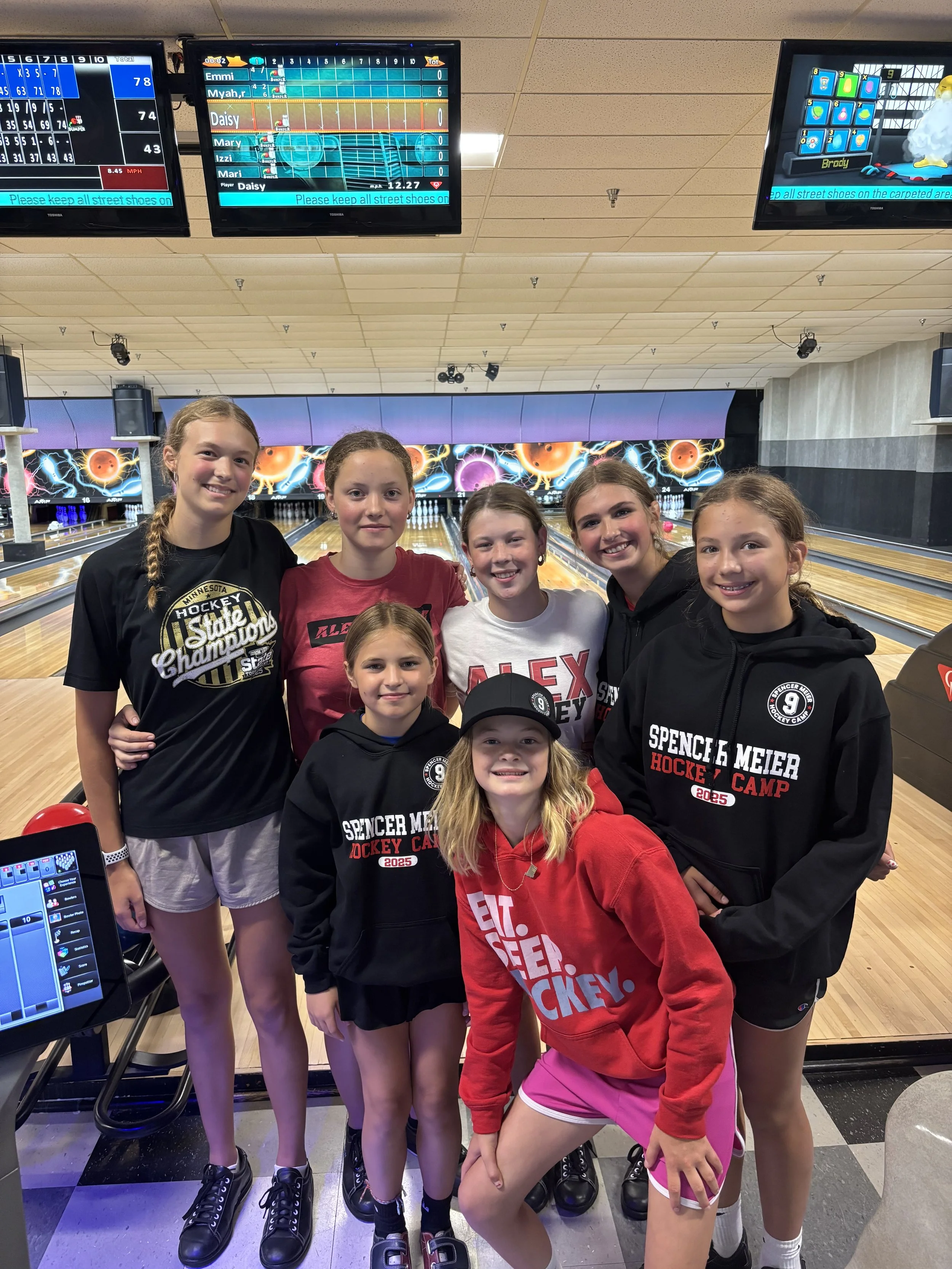 Group of seven young girls at a bowling alley, some wearing black 'Spencer Meier Hockey Camp' hoodies, one girl in a red hoodie with 'EAT SLEEP HOCKEY' text, and others in casual sportswear, standing together on the bowling lanes with digital scoreboards overhead.