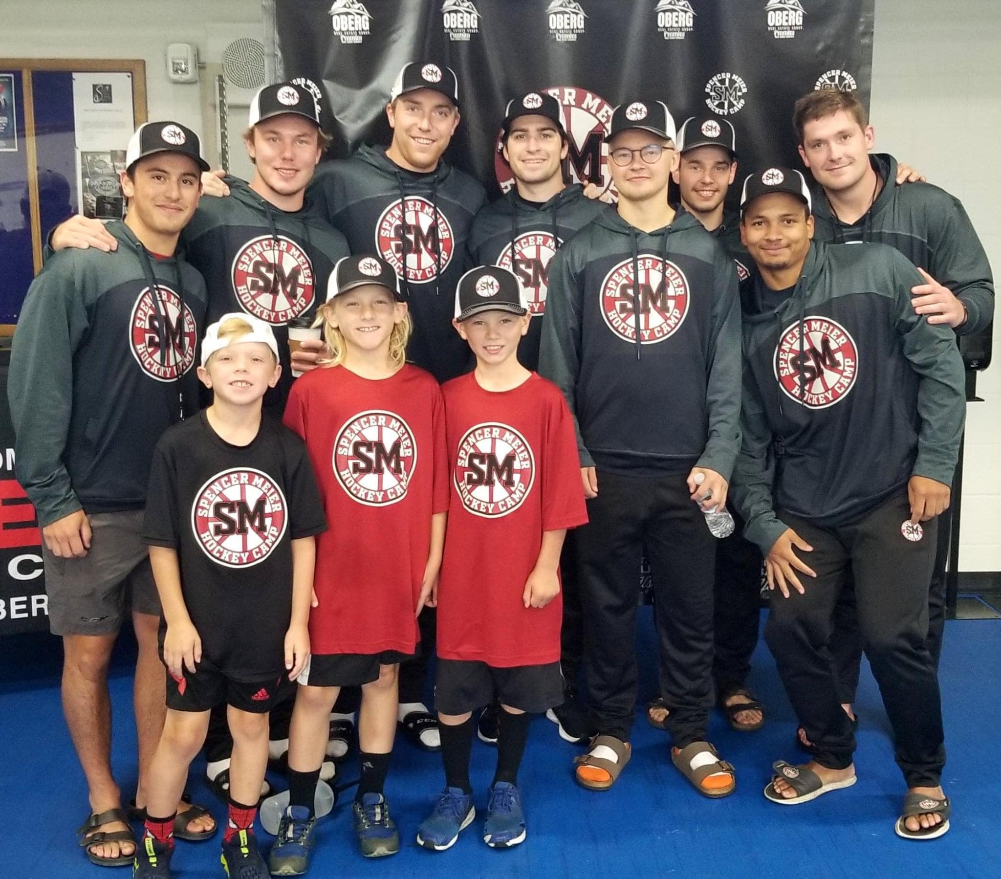 A group of people, including children and adults, posing together for a photo at a hockey camp. They are wearing hoodies, t-shirts, and hats with a logo that reads "Spencer Meier Hockey Camp" along with the initials "SM." Some are holding water bottles and all are smiling.