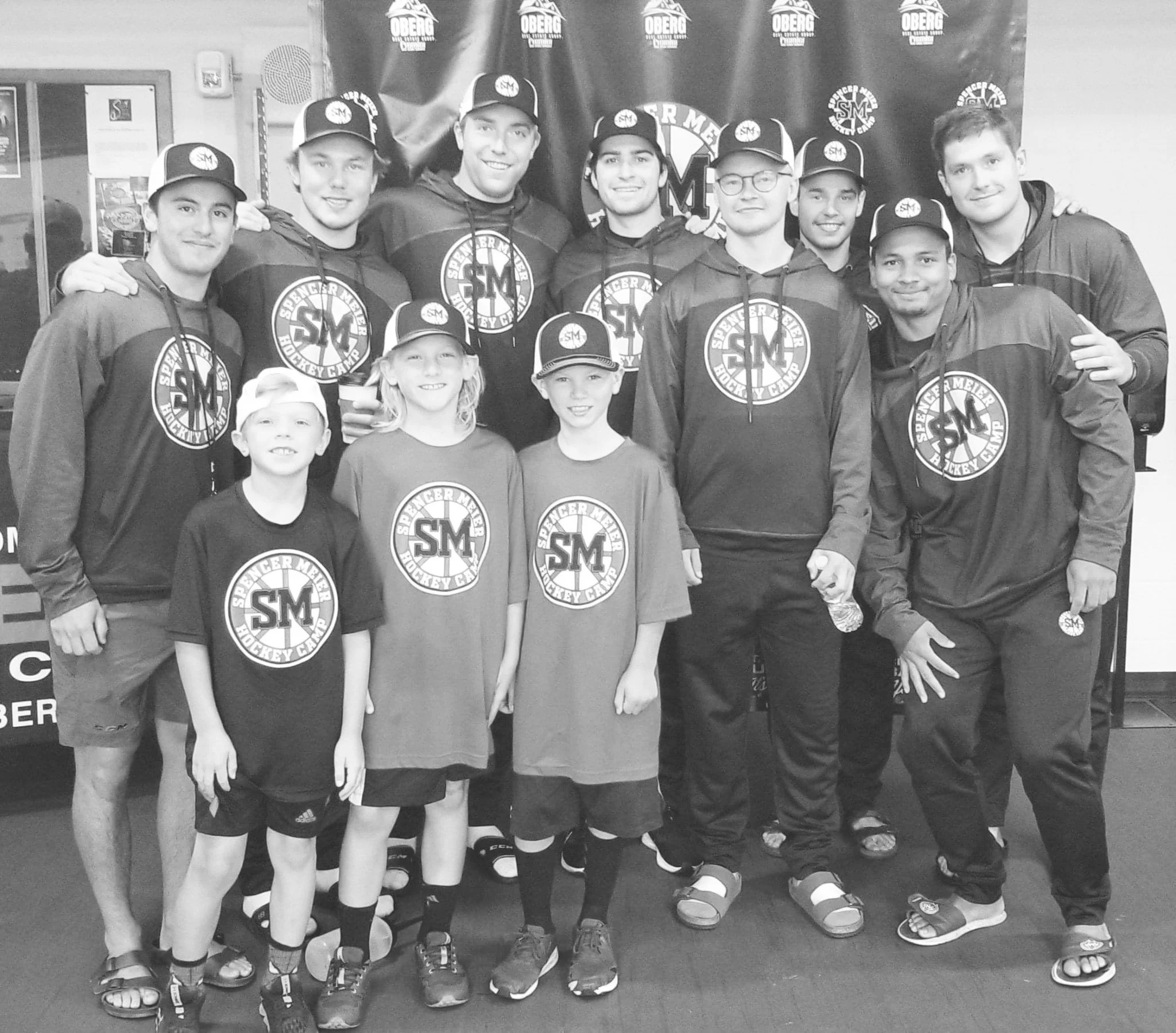 Group of eleven people, including children and adults, wearing hockey camp shirts and caps, posing together indoors in front of a banner that reads 'Spencer Mertens Hockey Camp'.