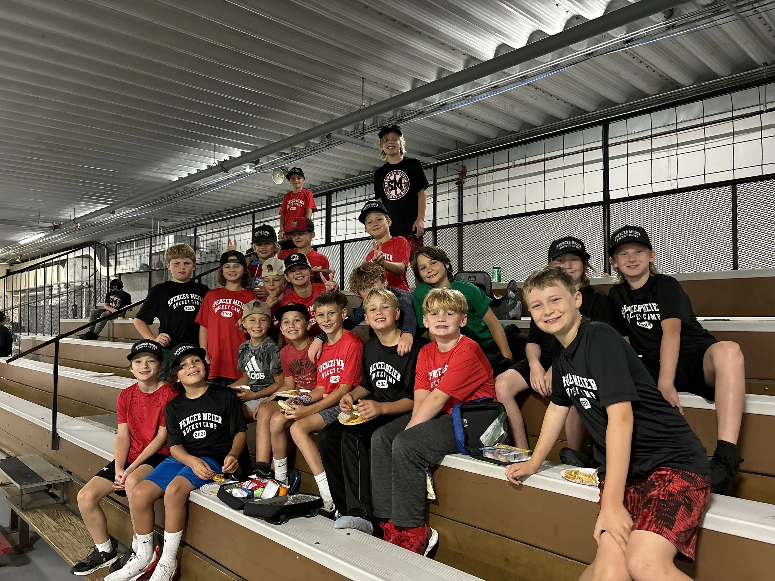 Group of young kids wearing hockey camp T-shirts, sitting on bleachers at an indoor ice rink.