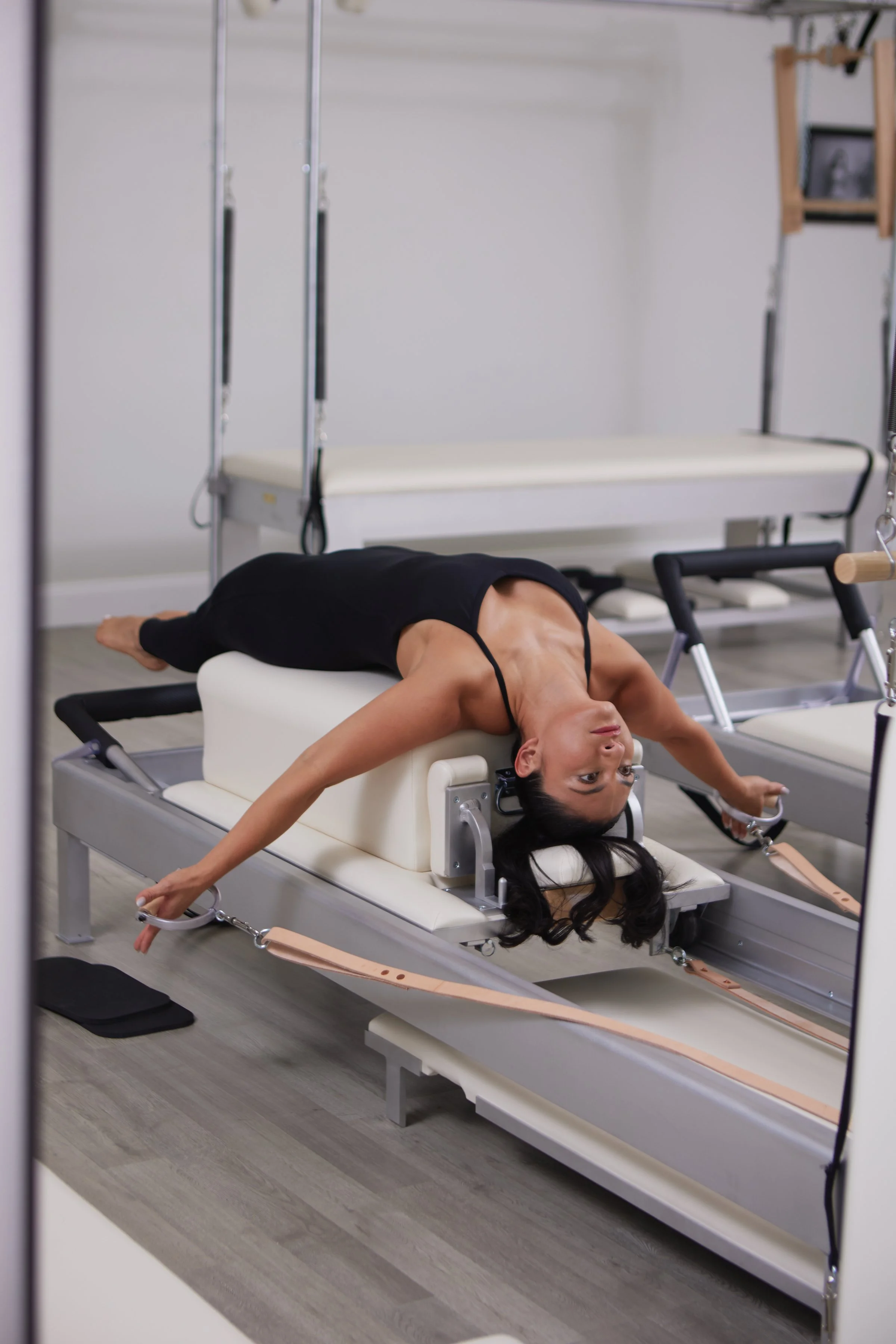 A woman is lying on a physical therapy rehab table, holding onto straps with her arms extended and her head upside down, in a rehabilitation or medical therapy room.