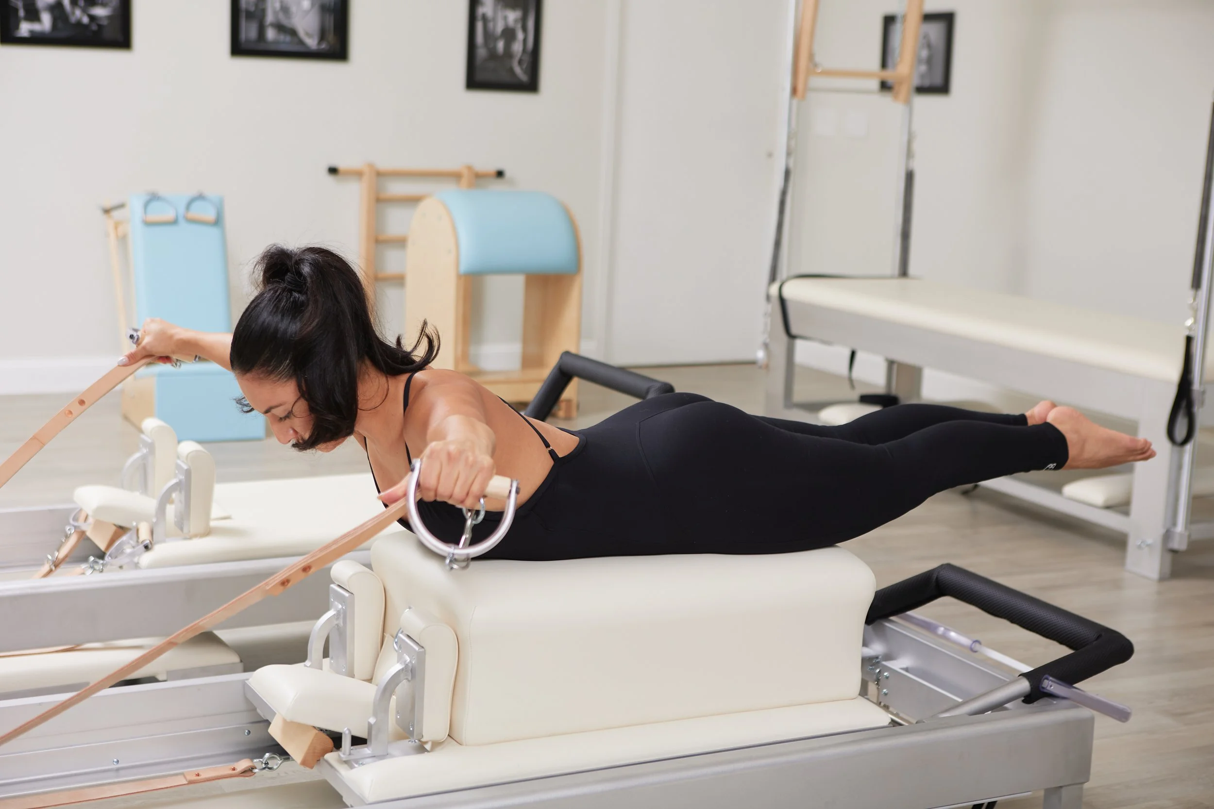 A woman with black hair in a ponytail performing a Pilates exercise on a reformer machine in a fitness studio, using resistance bands.
