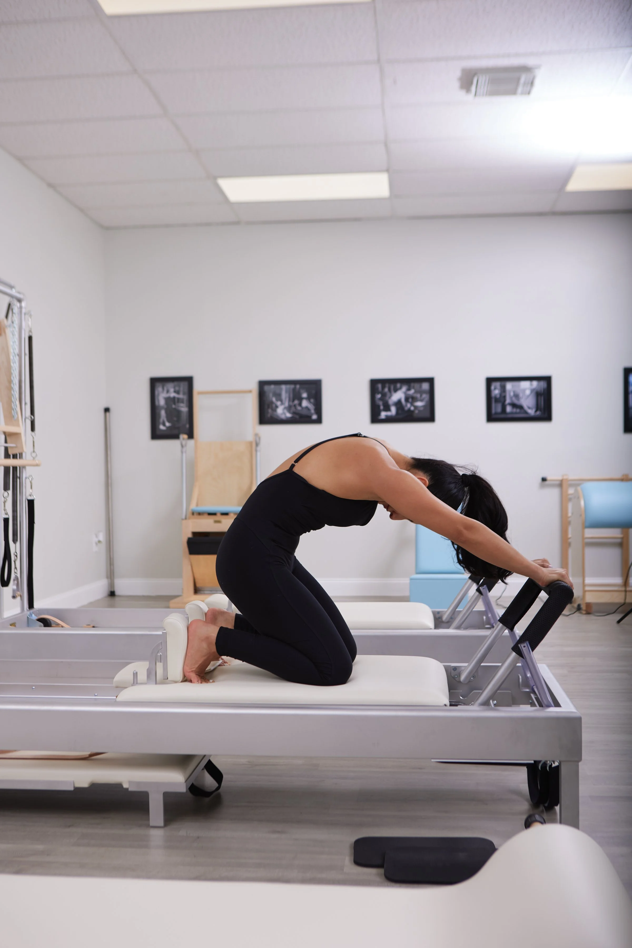 A woman in black workout clothes doing stretching exercises on specialized Pilates equipment in a fitness studio with framed photos on the wall.