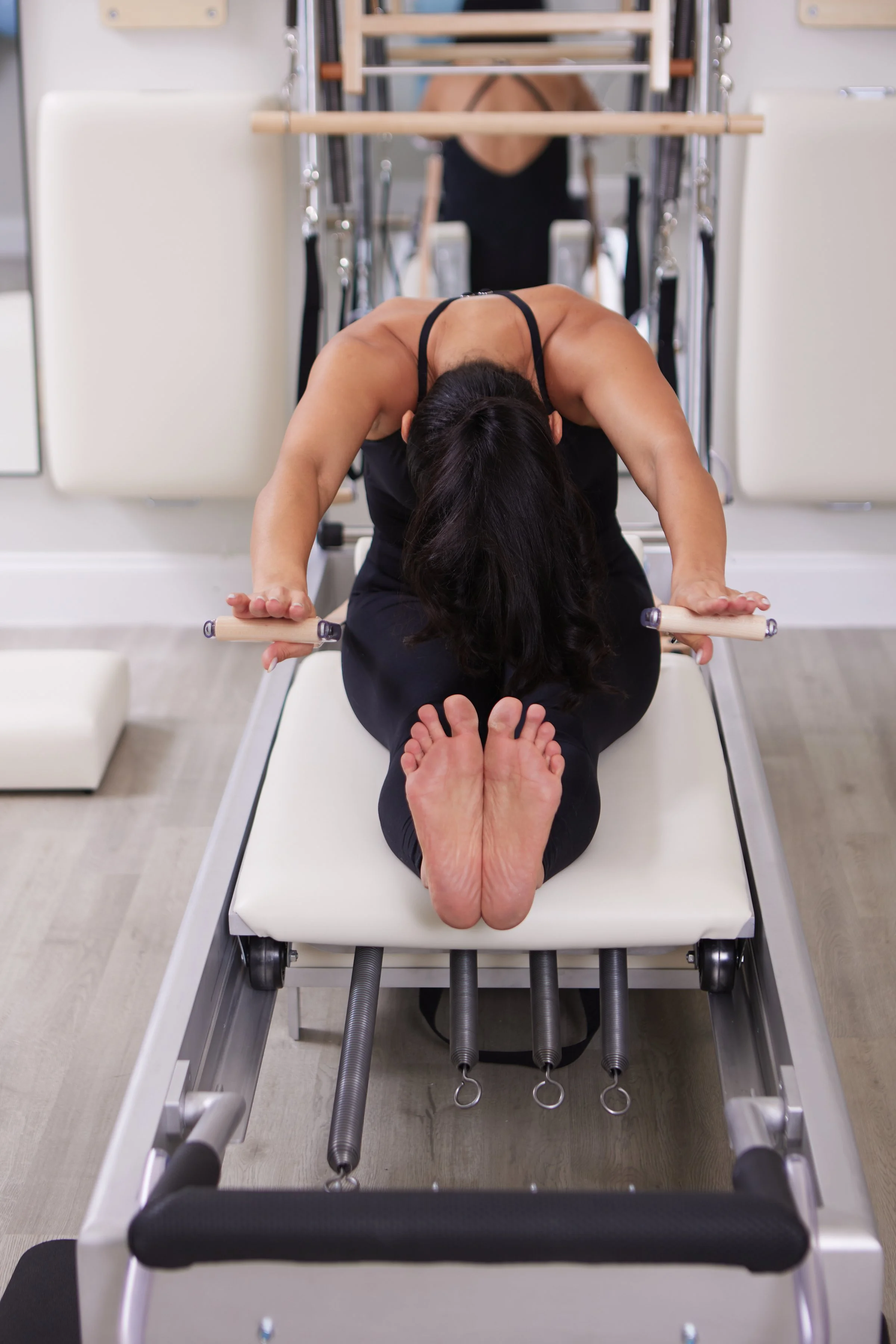 Woman in black athletic clothing performing a Pilates or yoga stretch on a Pilates reformer machine, with her body bent forward and arms extended, head resting on her knees.