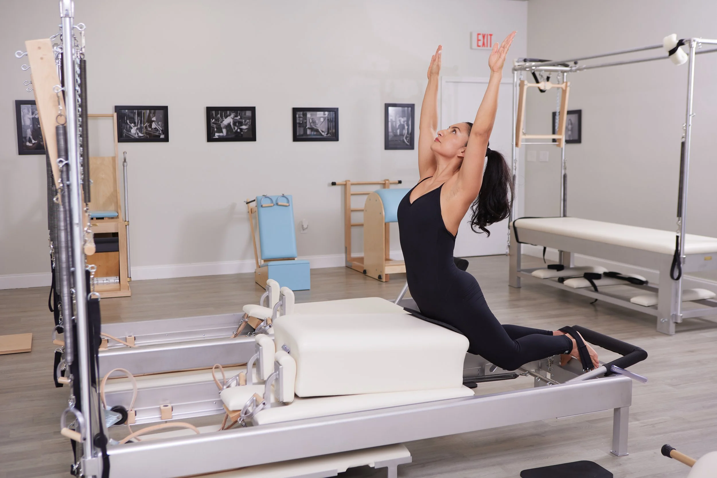 A woman in a black workout outfit performing a stretch on Pilates reformer equipment in a Pilates studio with framed black-and-white photos on the wall.