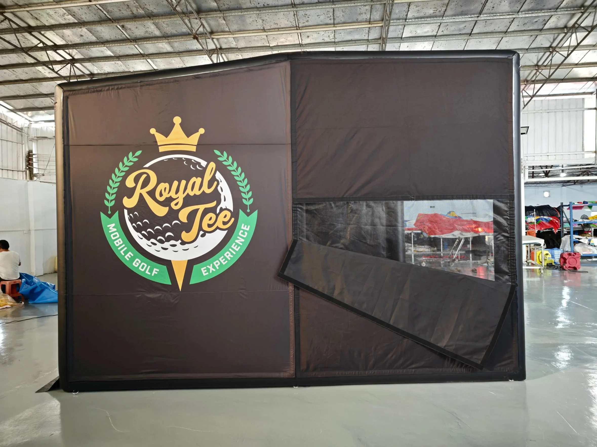 Black portable golf simulator with a logo that reads 'Royal Tee Mobile Golf Experience,' featuring a crown, golf ball, and wreath, inside a spacious warehouse