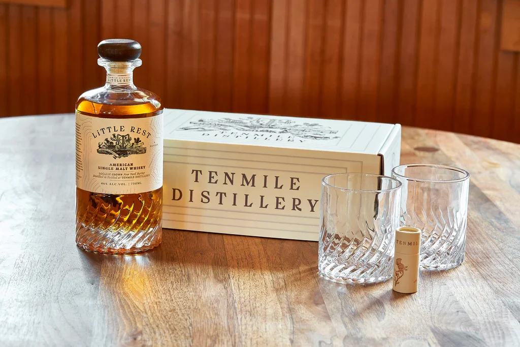 A bottle of Little Rest American single malt whisky, two empty glasses, a small cylindrical container, and a Tenmile Distillery box on a wooden table.