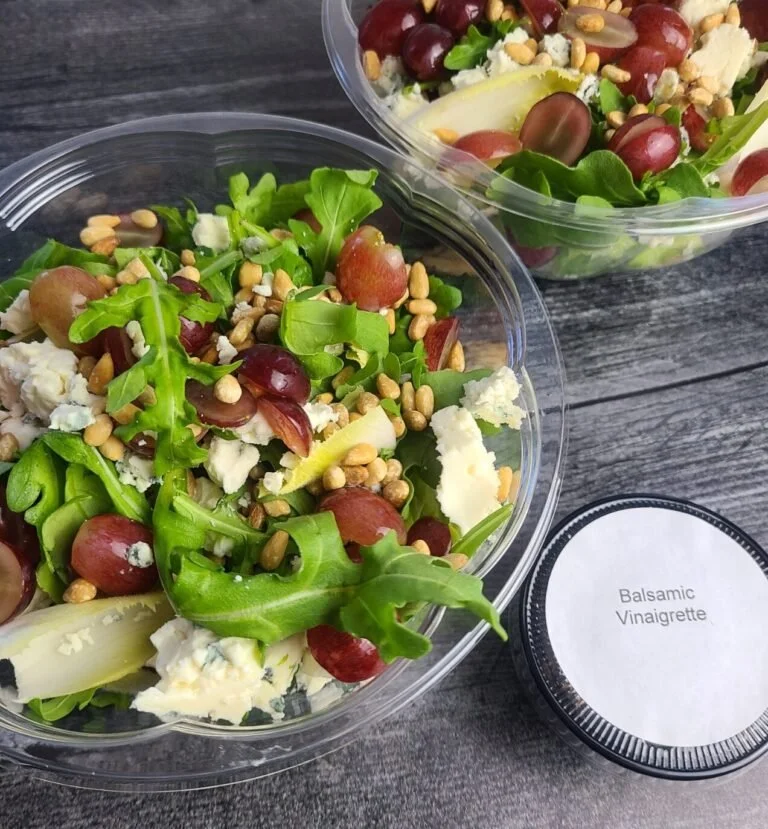 Two bowls of mixed green salads with grapes, cheese, pine nuts, and lettuce, alongside a container labeled Balsamic Vinaigrette.