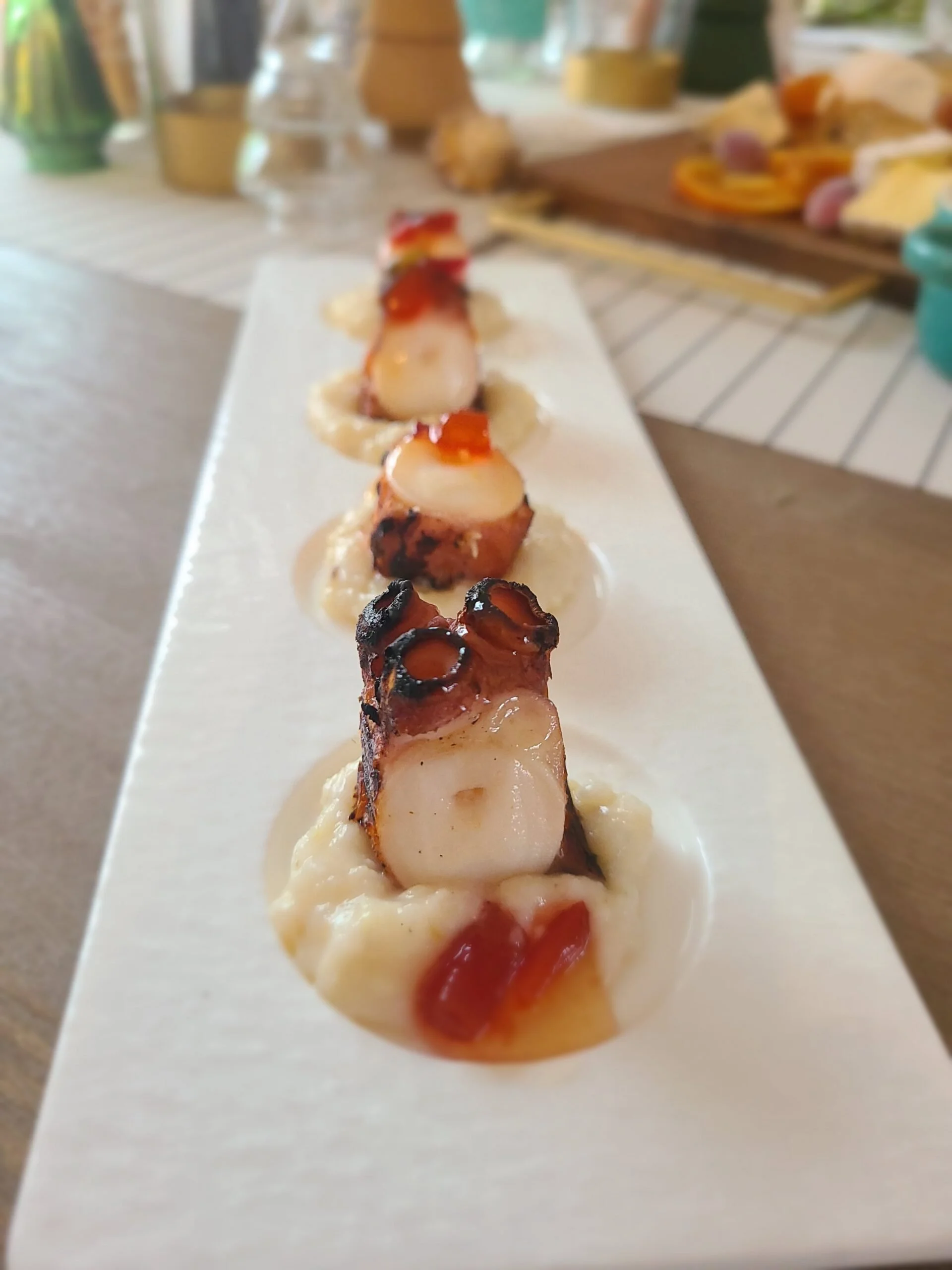 Close-up of a row of small appetizer bites on a white rectangular plate, topped with different sauces and garnishes, in a bright, airy kitchen setting.
