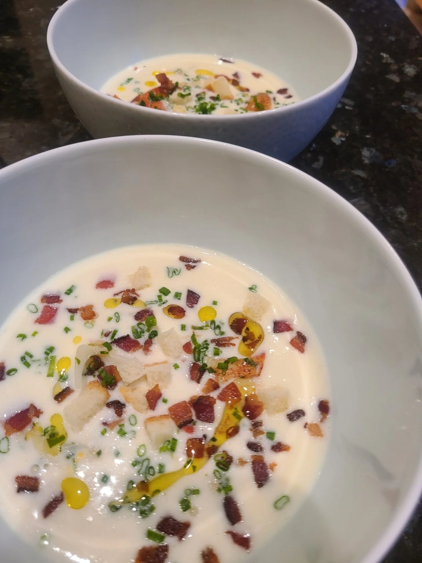 Bowl of creamy soup garnished with chopped chives, bacon bits, and croutons on a dark granite countertop.