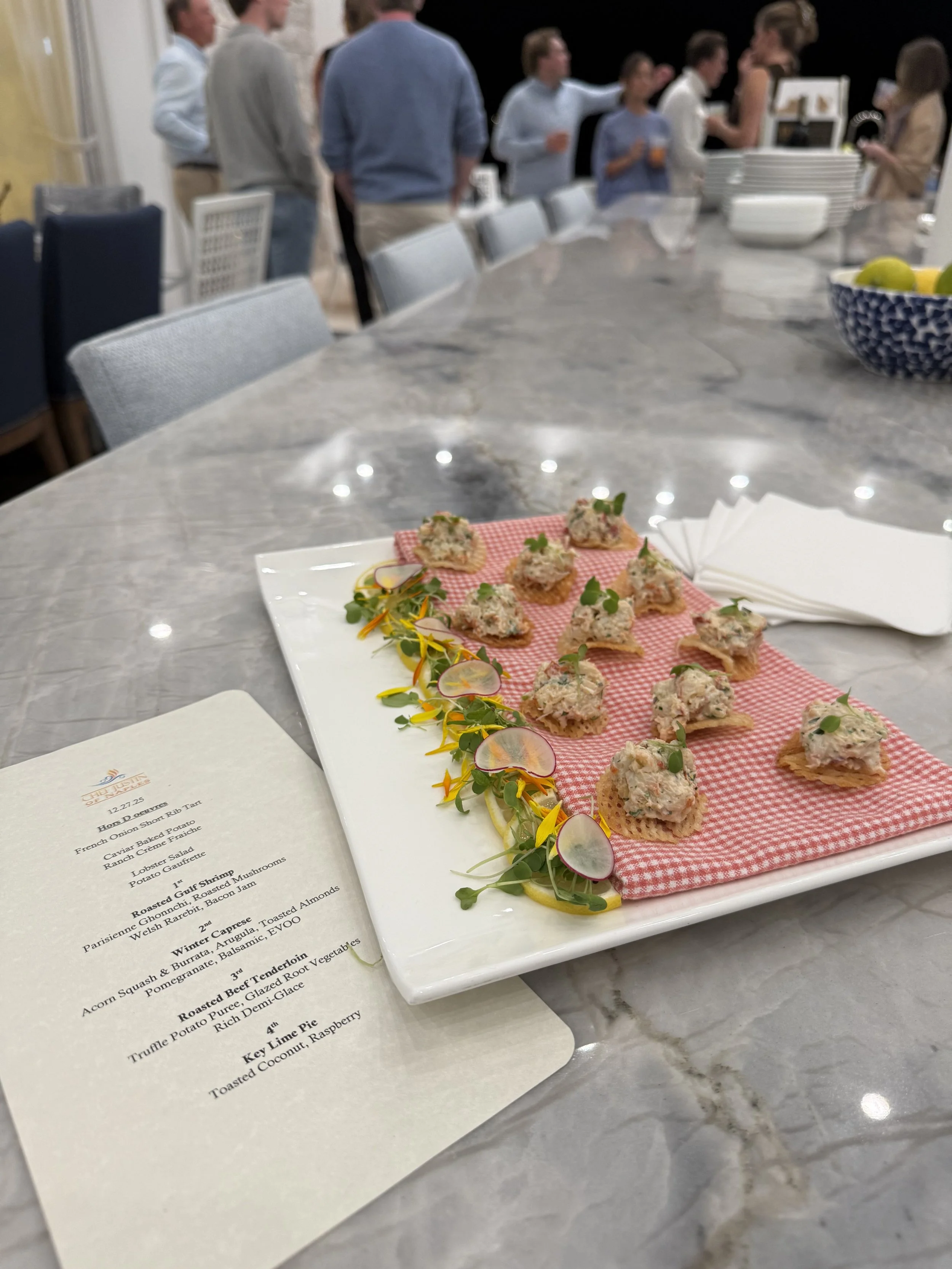 A marble table with a menu and a white rectangular platter holding small appetizers garnished with herbs and radish slices, with a group of people in the background at a social gathering.
