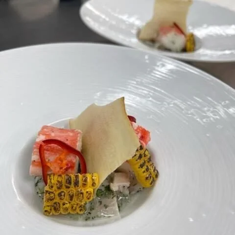 A plated dish featuring grilled corn on the cob, seafood, and garnishes on a white plate.
