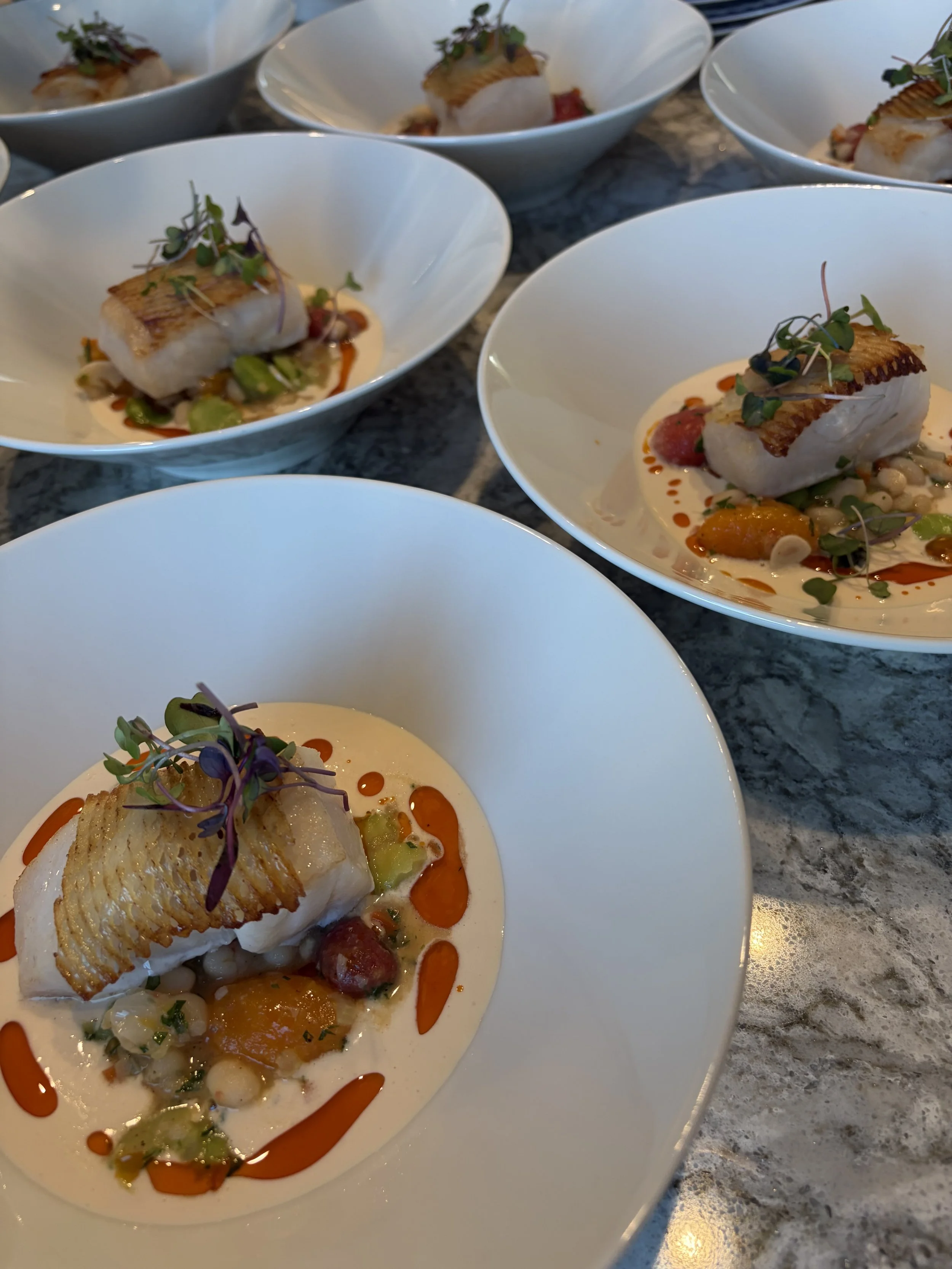 Crispy fish fillet served over a bed of vegetables with sauce and microgreens in white bowls on a marble surface.