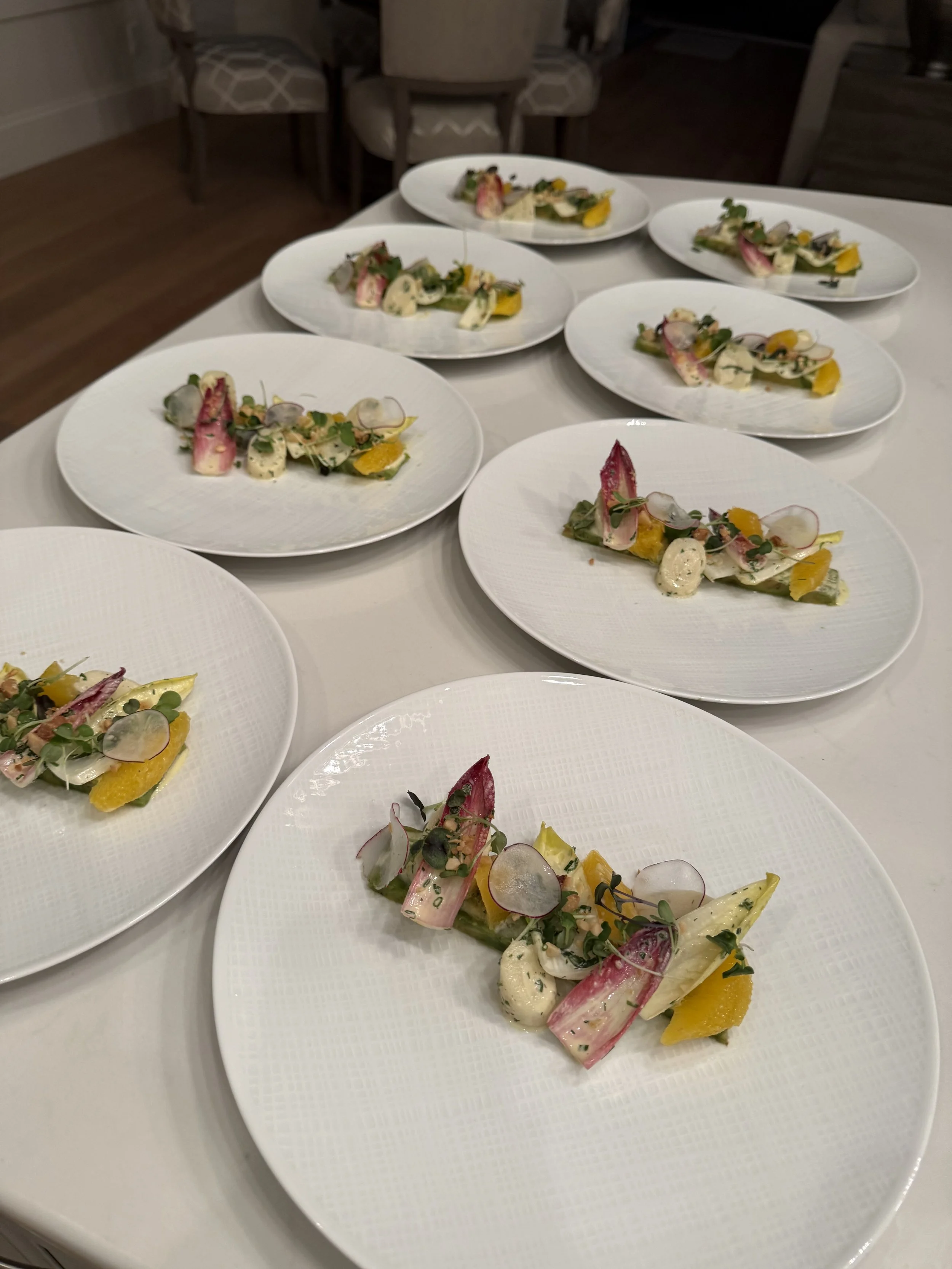 Multiple white plates with gourmet salads featuring radish slices, microgreens, edible flowers, yellow and green vegetables, and creamy dollops, arranged on a white table.