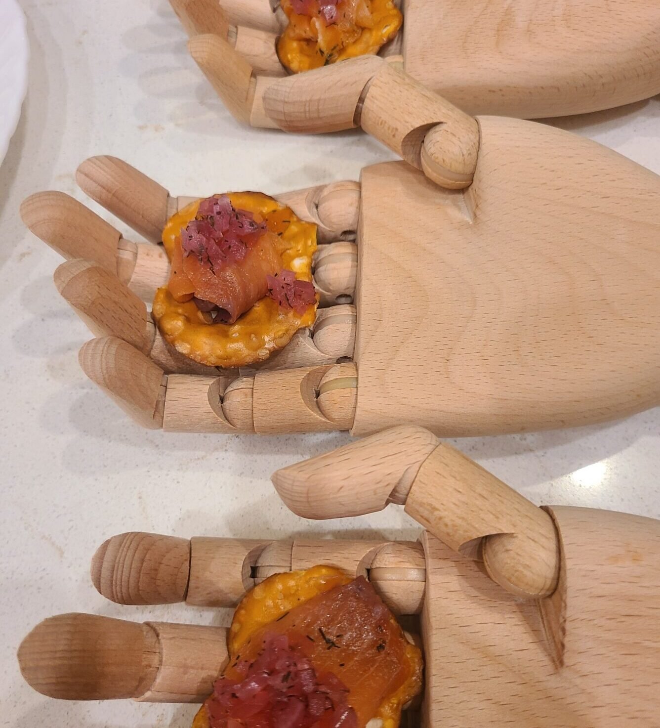 Multiple wooden hand models with sandwiches on top, each sandwich has cheese, ham, and pickled vegetables, on a light-colored countertop.