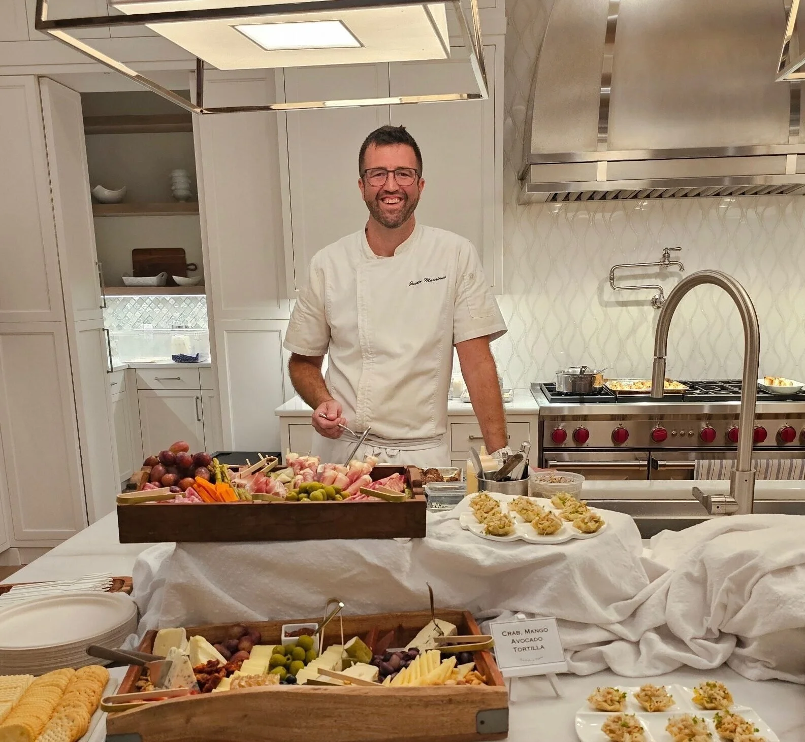 Chef standing in a kitchen behind a table filled with various food items, including a cheese platter, grapes, and appetizers.