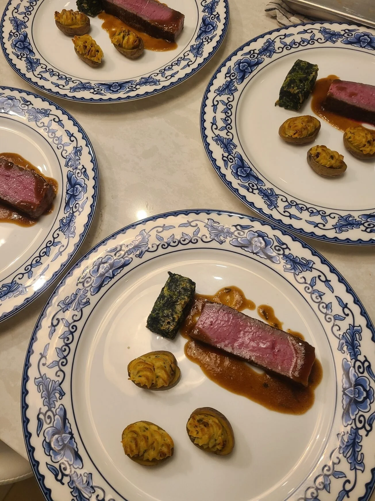 Multiple white plates with blue floral patterns, each containing a piece of cooked steak with sauce, roasted vegetables, and a vegetable roll.
